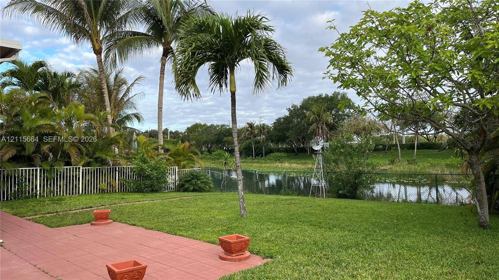 19409 NW 24th Place, Pembroke Pines, FL 33029 Rear exterior featuring a paved terrace, expansive lawn, mature palm trees, metal fencing, and a waterfront backdrop with a decorative windmill