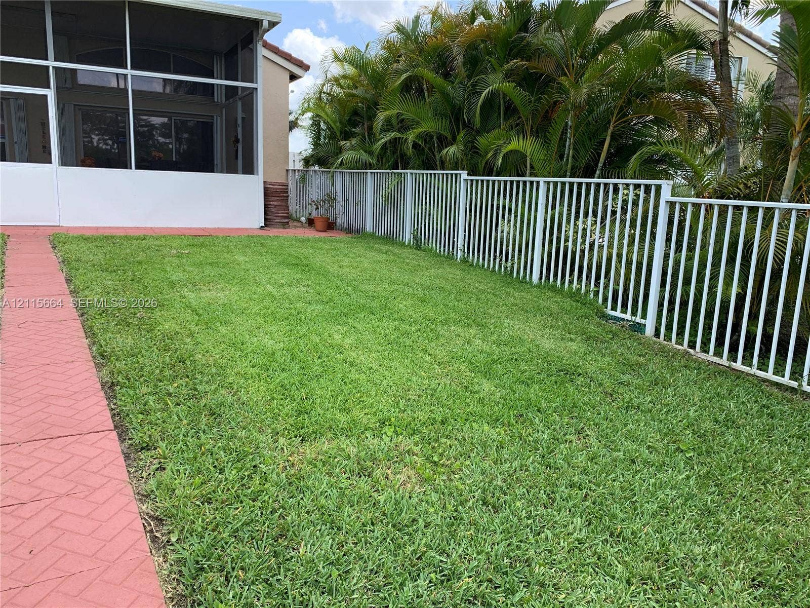19409 NW 24th Place, Pembroke Pines, FL 33029 Backyard featuring a grass lawn, white metal fencing, a red brick-pattern paver walkway, and a screened-in porch area