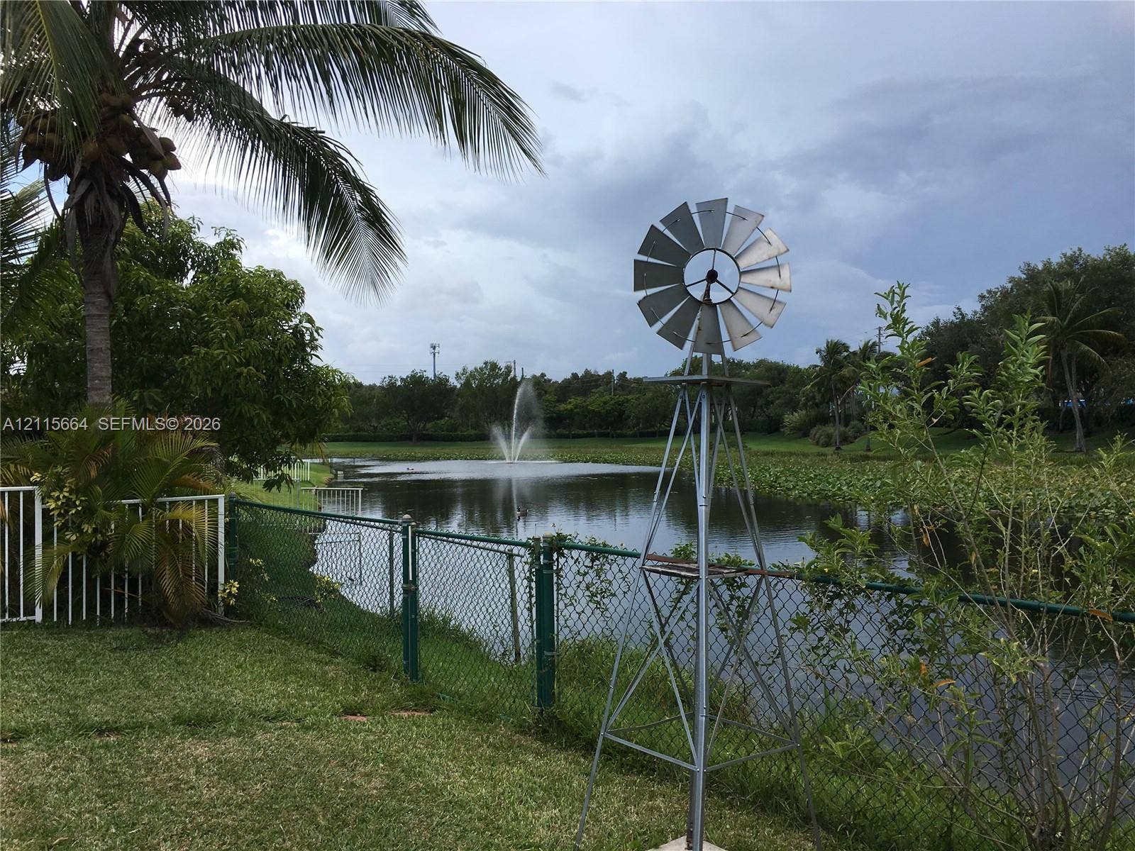 19409 NW 24th Place, Pembroke Pines, FL 33029 Waterfront grounds featuring a metal windmill, a center-pond fountain, chain-link perimeter fencing, and mature palm trees