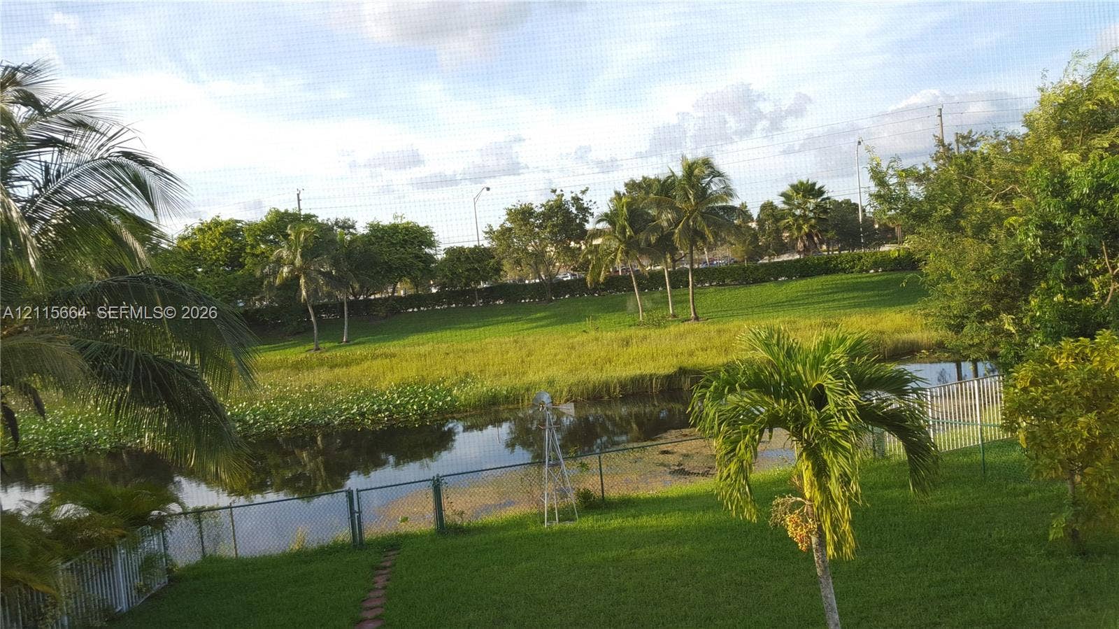 19409 NW 24th Place, Pembroke Pines, FL 33029 Private backyard featuring a grassy lawn, mature palm trees, a decorative metal windmill, and water frontage with a metal fence enclosure