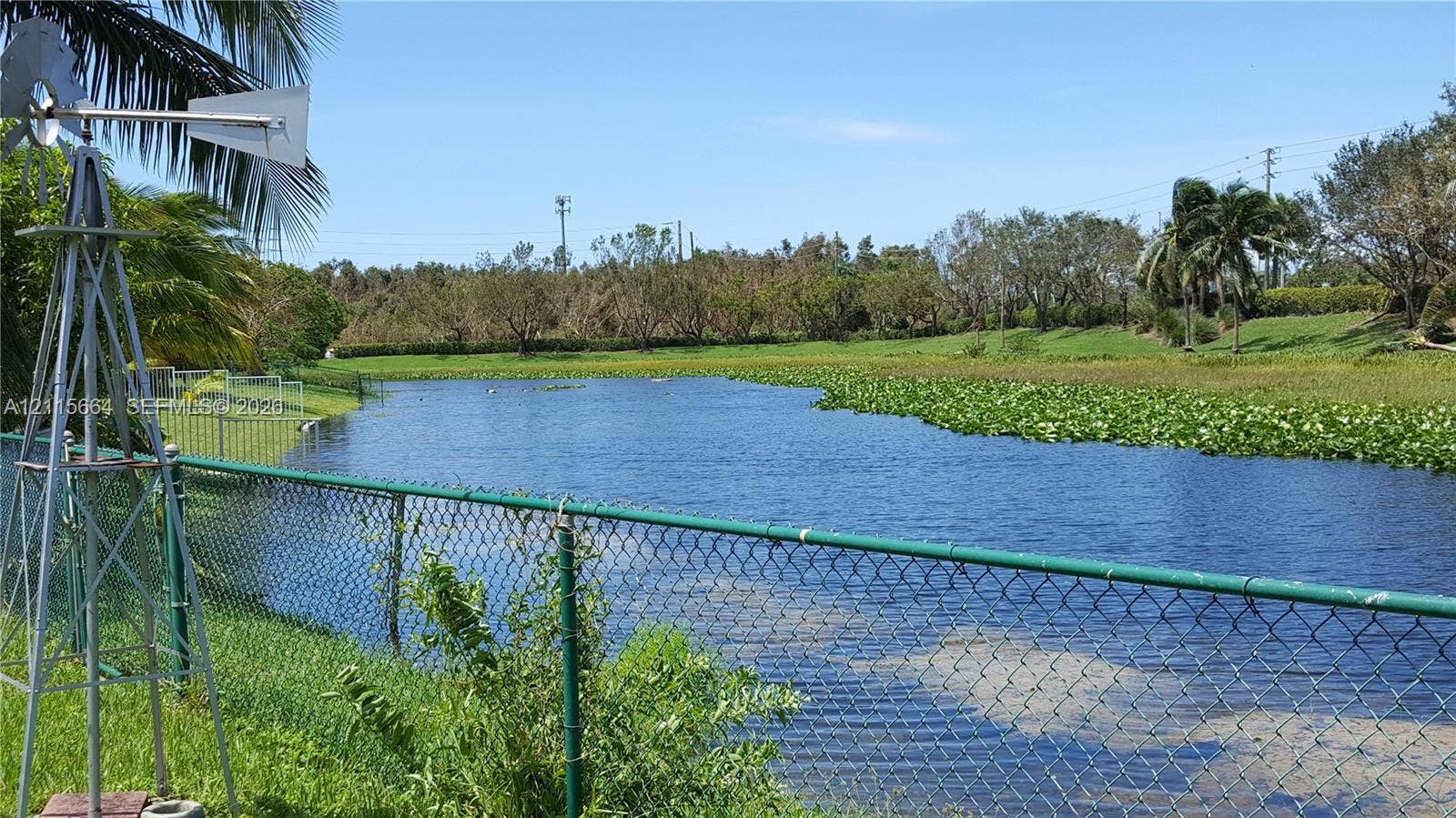 19409 NW 24th Place, Pembroke Pines, FL 33029 Waterfront lot featuring a metallic windmill, green chain-link perimeter fencing, and mature vegetation
