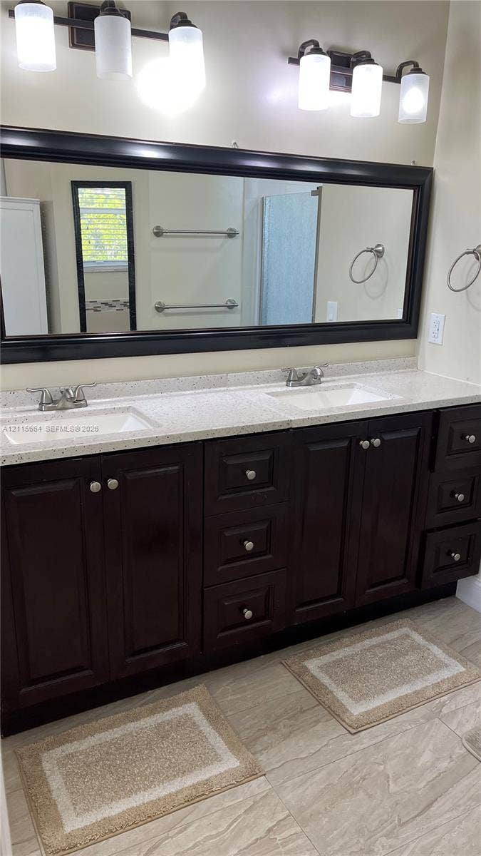 19409 NW 24th Place, Pembroke Pines, FL 33029 Dual-sink vanity featuring dark wood cabinetry, speckled stone-composite countertops, large framed mirror, and two multi-bulb overhead light fixtures