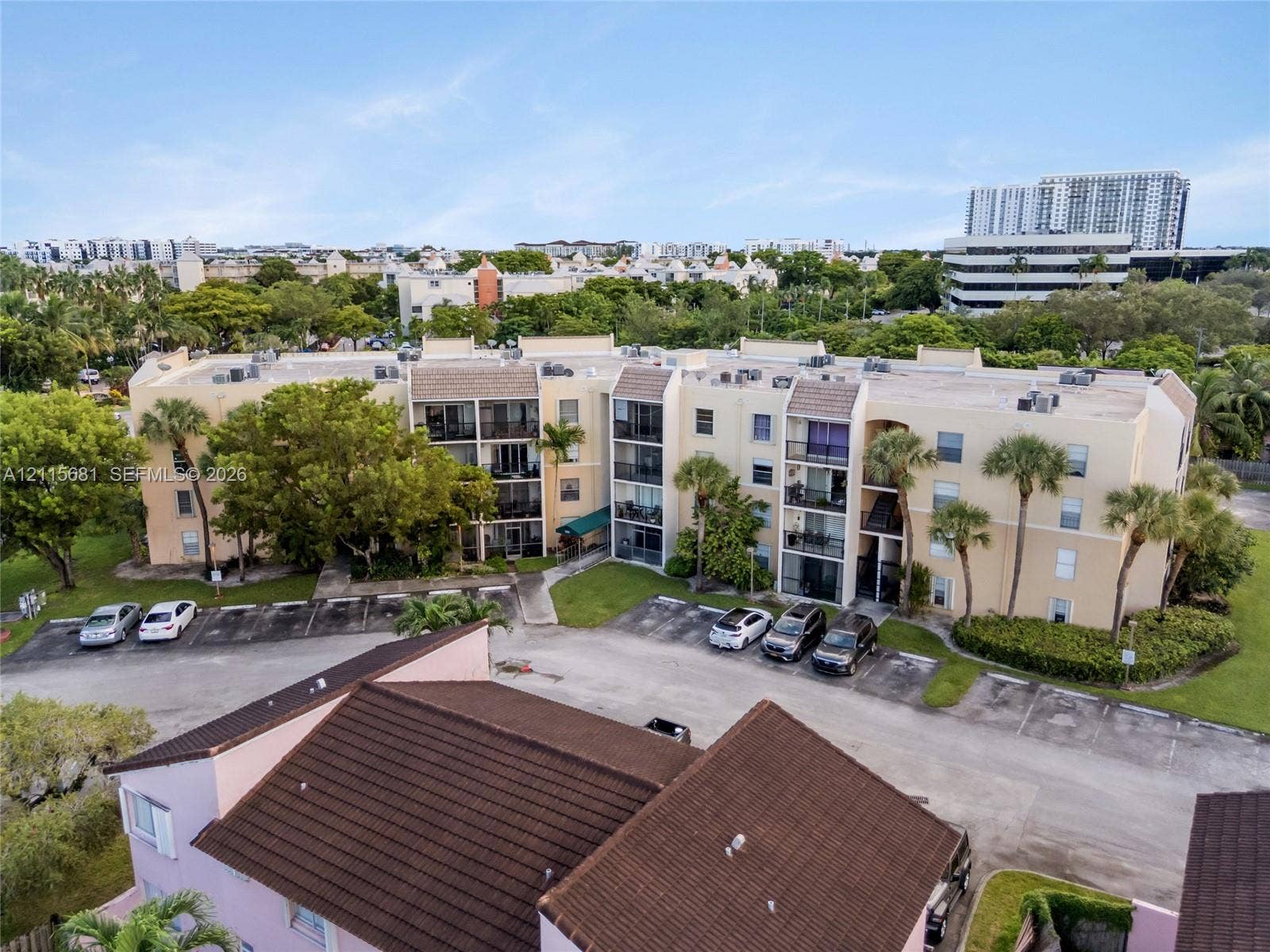 Multi-story residential building featuring a beige stucco exterior, tiled roofing, private balconies, and dedicated asphalt parking areas surrounded by mature palm trees and green space