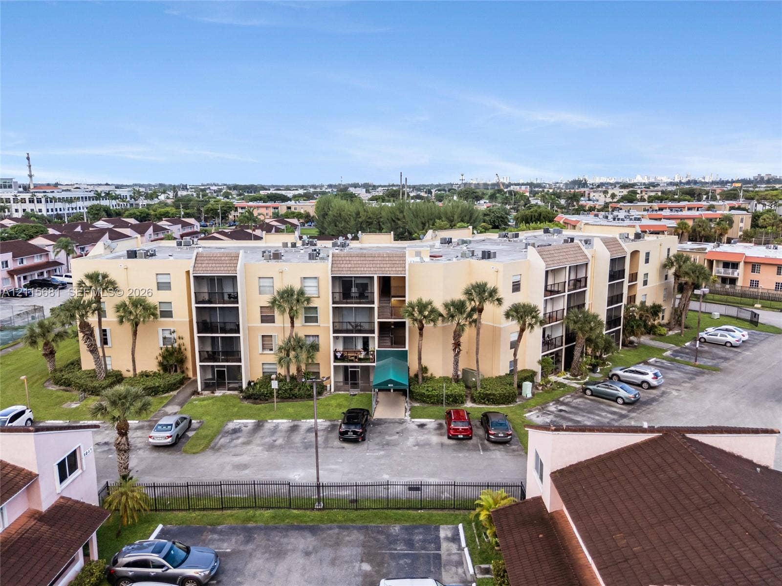 Multi-story residential building exterior featuring a stucco facade, integrated screened-in balconies, a shingled roof, on-site surface parking, and mature palm tree landscaping