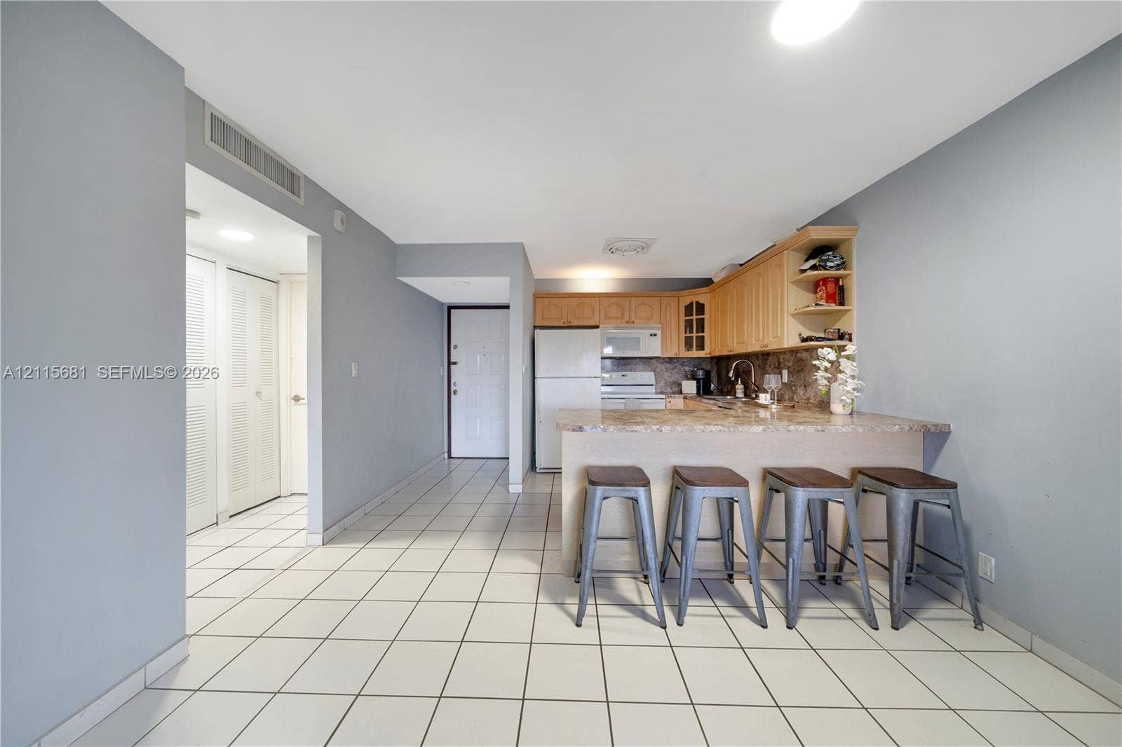 Kitchen area featuring wood-finish cabinetry, stone-patterned countertops, integrated breakfast bar, white appliance suite, and neutral tile flooring