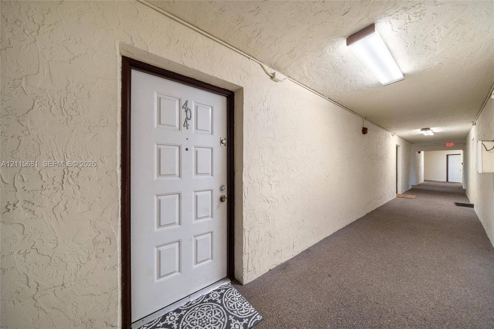 White six-panel entry door with integrated peephole and metallic hardware, textured wall finishes, and overhead surface-mounted lighting