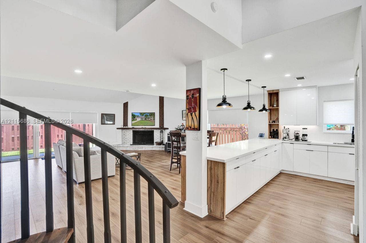 8946 SW 150th Ct Circle E, Miami, Miami, FL 33196 Open-concept kitchen featuring high-gloss white cabinetry, expansive countertop peninsula, wood-finish flooring, and industrial-style pendant lighting, complemented by a floor-to-ceiling brick fireplace