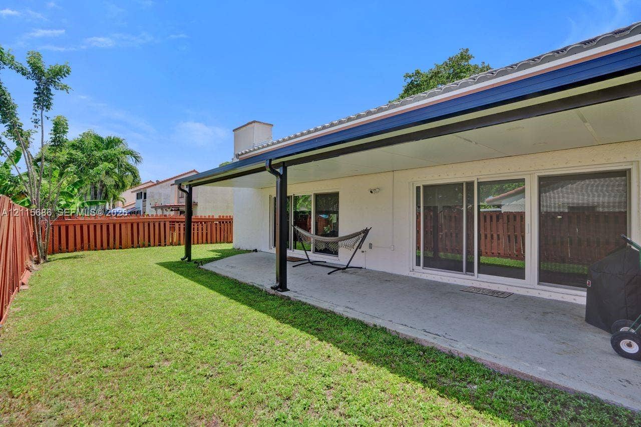 8946 SW 150th Ct Circle E, Miami, Miami, FL 33196 Covered concrete patio with metal support columns, sliding glass doors, and perimeter wood fencing surrounding a grass lawn