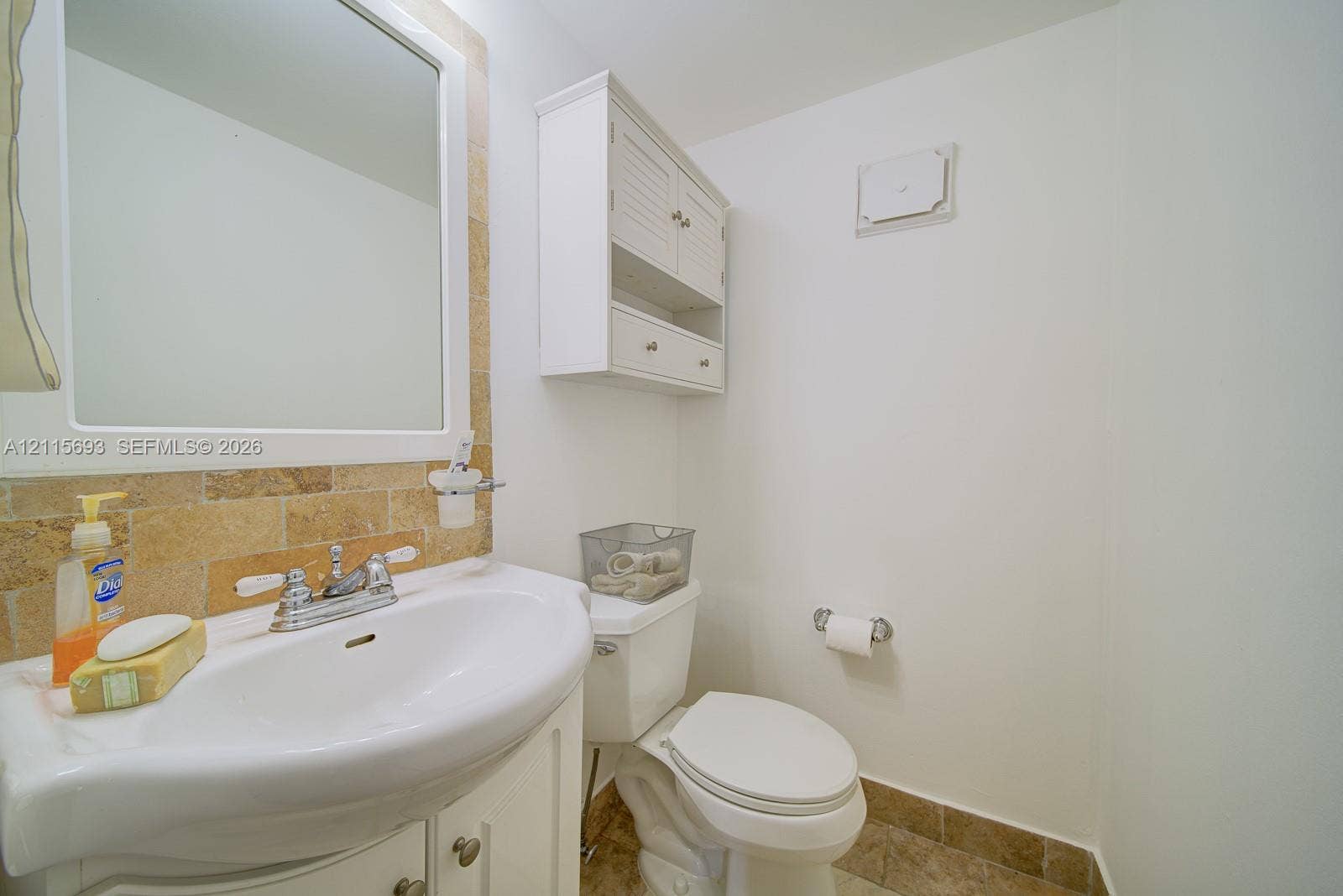 Bathroom featuring a white vanity cabinet with a pedestal sink, wall-mounted white cabinetry, stone-textured tile backsplash, and stone-finish flooring