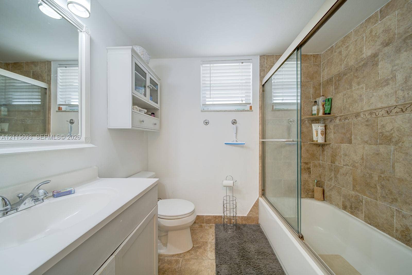 Bathroom featuring a white vanity cabinet with an integrated sink, wall-mounted cabinetry, a glass-enclosed bathtub shower combination, and stone-finish tile surround
