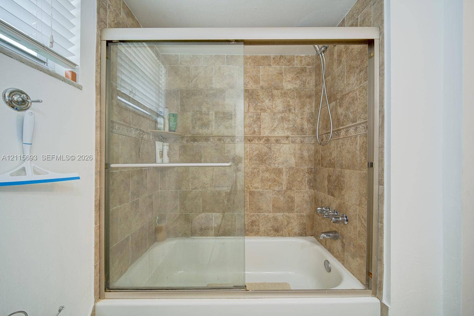 Shower and bathtub combination featuring a sliding glass enclosure, floor-to-ceiling textured tile surround, wall-mounted handheld showerhead, and integrated soap niche