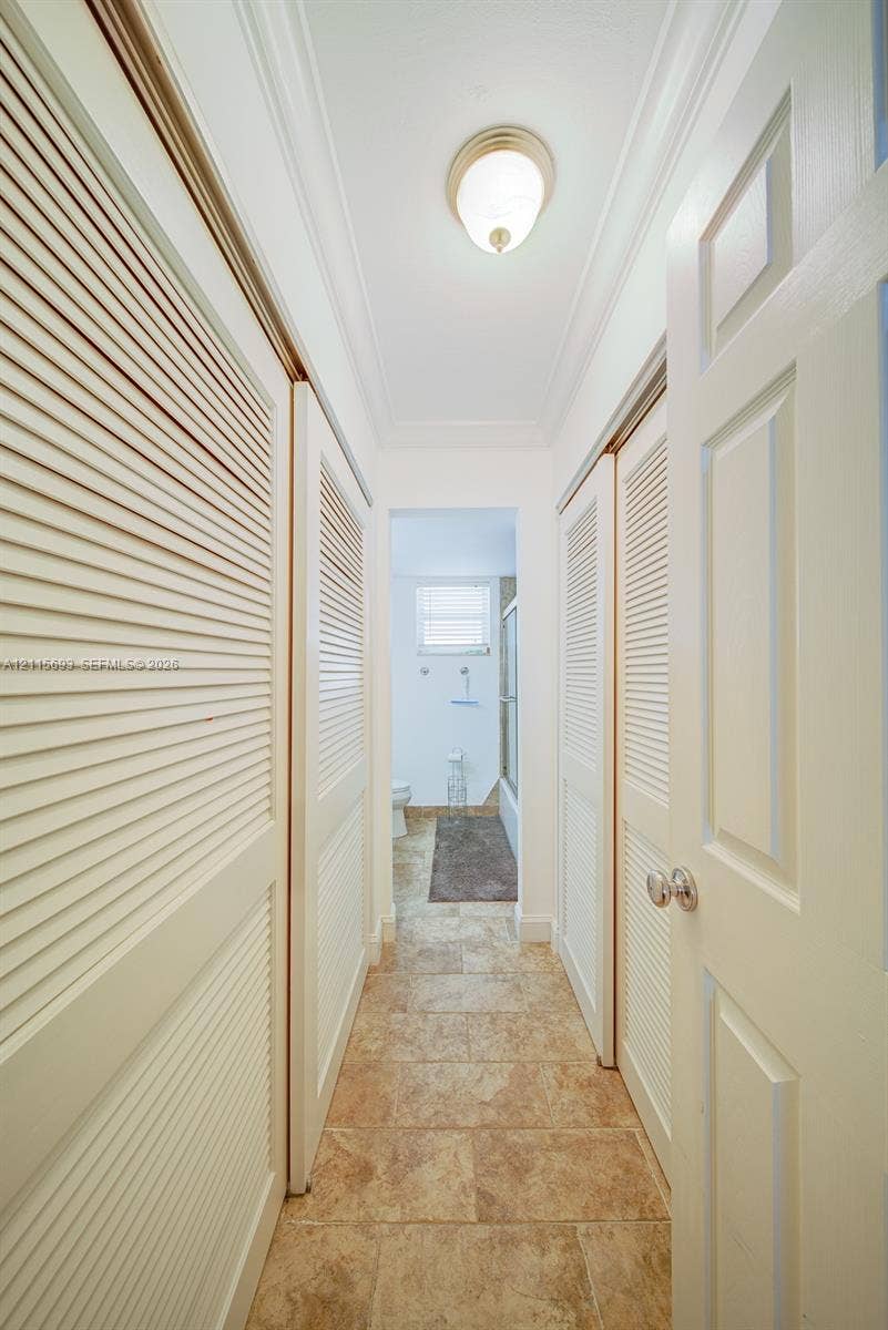 Hallway featuring dual sets of white louvered doors, crown molding, stone-finish tile flooring, and a flush-mount light fixture