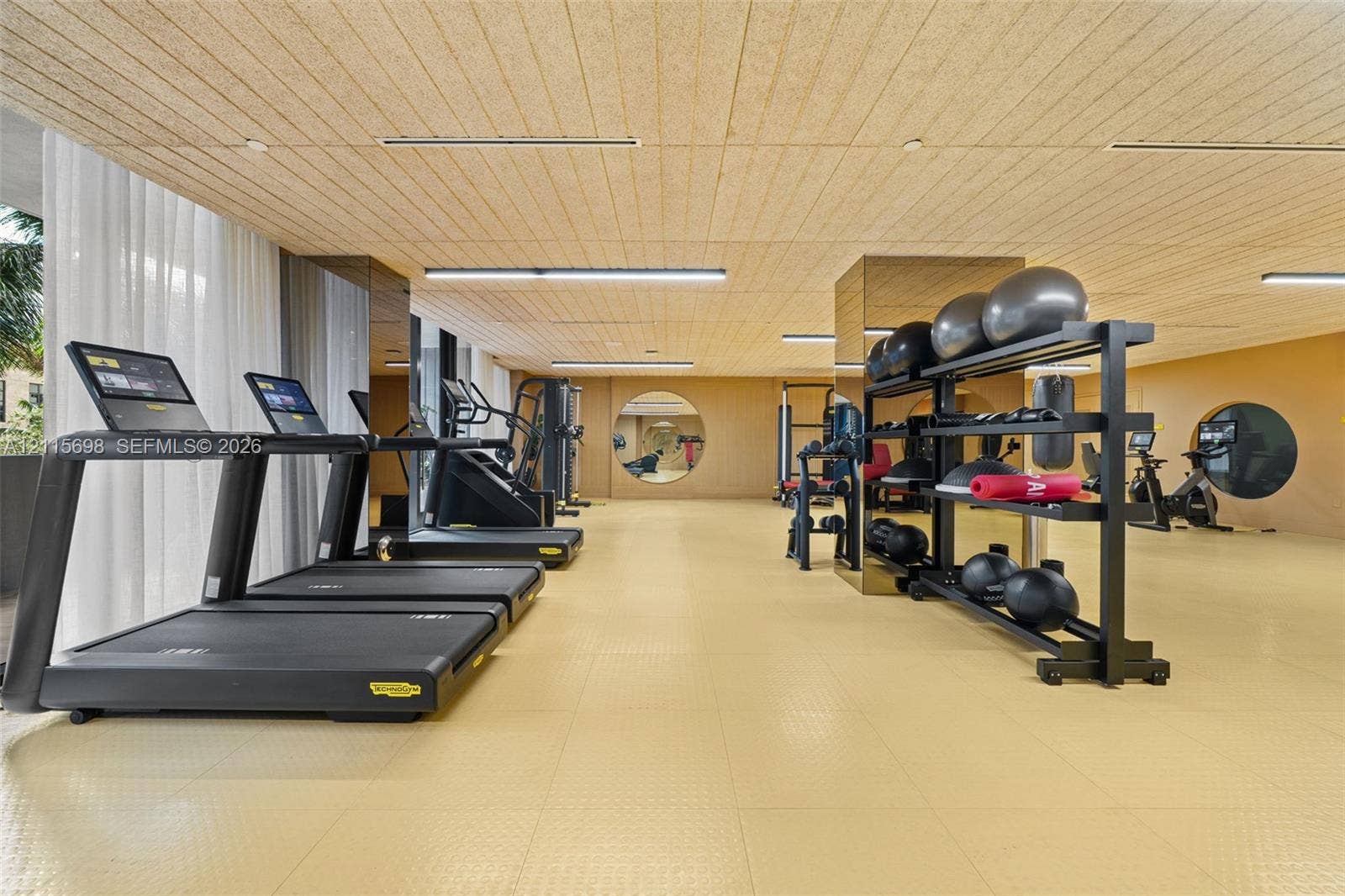 Fitness area featuring textured wood-panel ceiling, mirrored structural pillars, circular accent mirrors, recessed linear lighting, and resilient athletic flooring