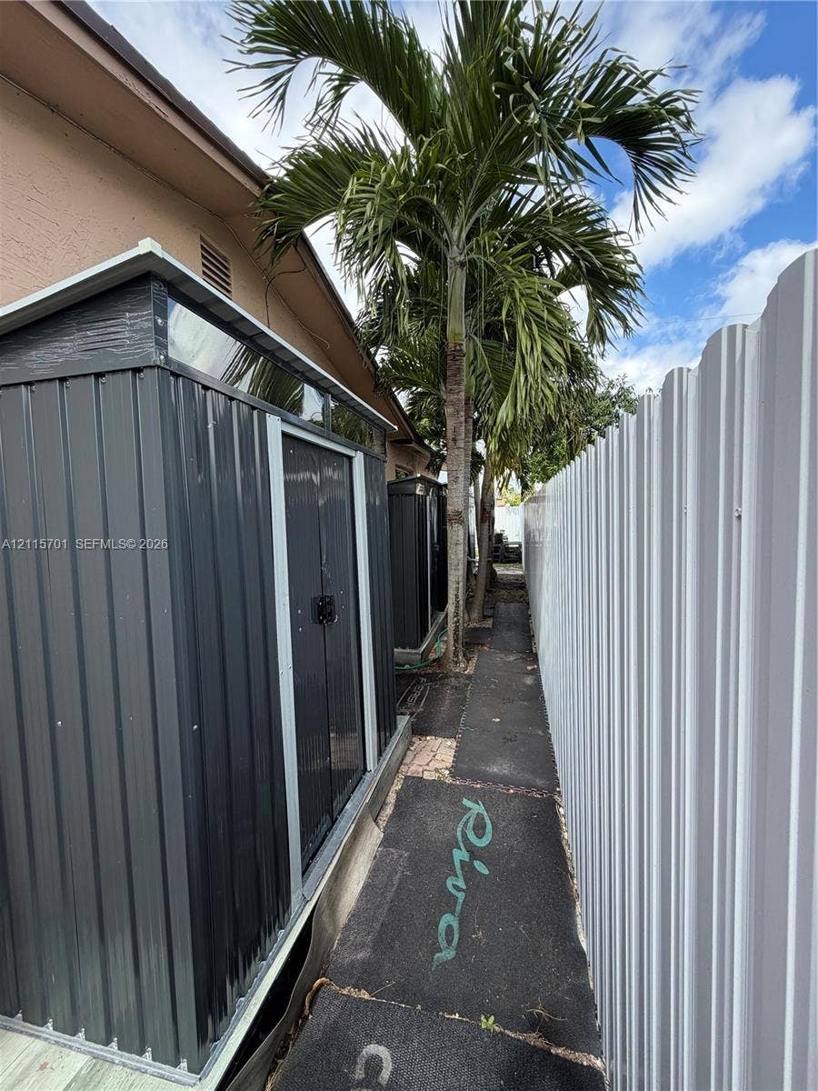 8641 NW 25th Court, #864, Sunrise, Sunrise, FL 33322 Metal storage structure featuring a gable roof, corrugated siding, and double doors, situated along a paved exterior pathway bordered by a metal privacy fence