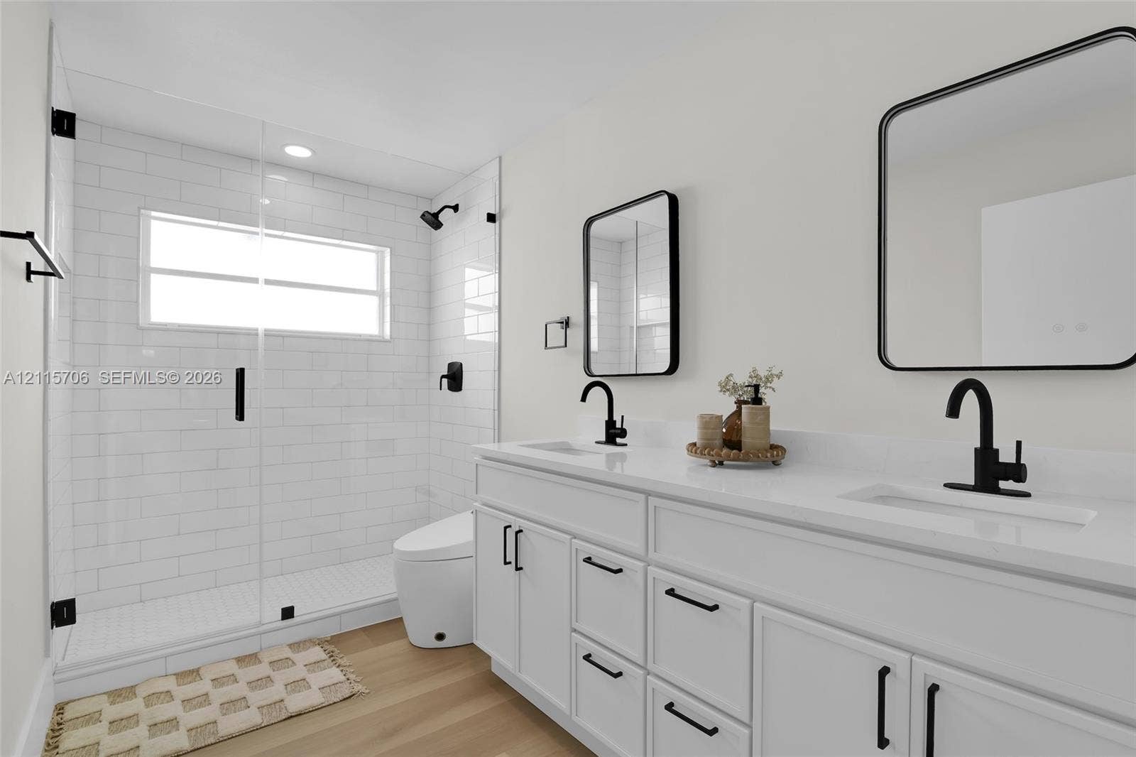 5184 Deerhurst Crescent Circle, Boca Raton, FL 33486 Dual-vanity cabinetry with white surfaces and integrated rectangular sinks, accented by matte black fixtures, framed mirrors, a walk-in shower with glass enclosure, and wood-finish flooring