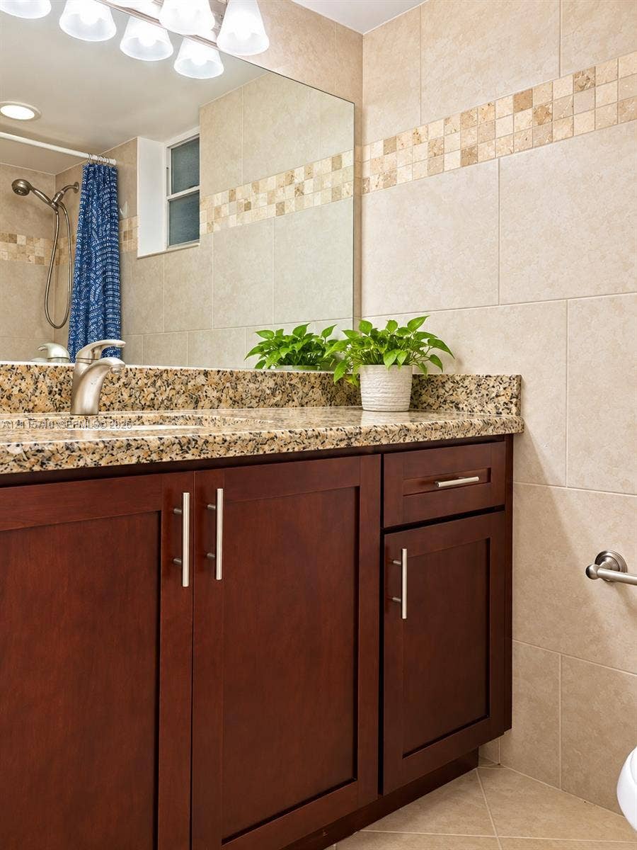 406 NW 68th Avenue, #115, Plantation, Plantation, FL 33317 Vanity featuring dark wood-finish cabinetry, stone countertop, undermount sink, and polished metal faucet hardware, complemented by floor-to-ceiling wall tile with mosaic inlay and an expansive wall-mounted mirror