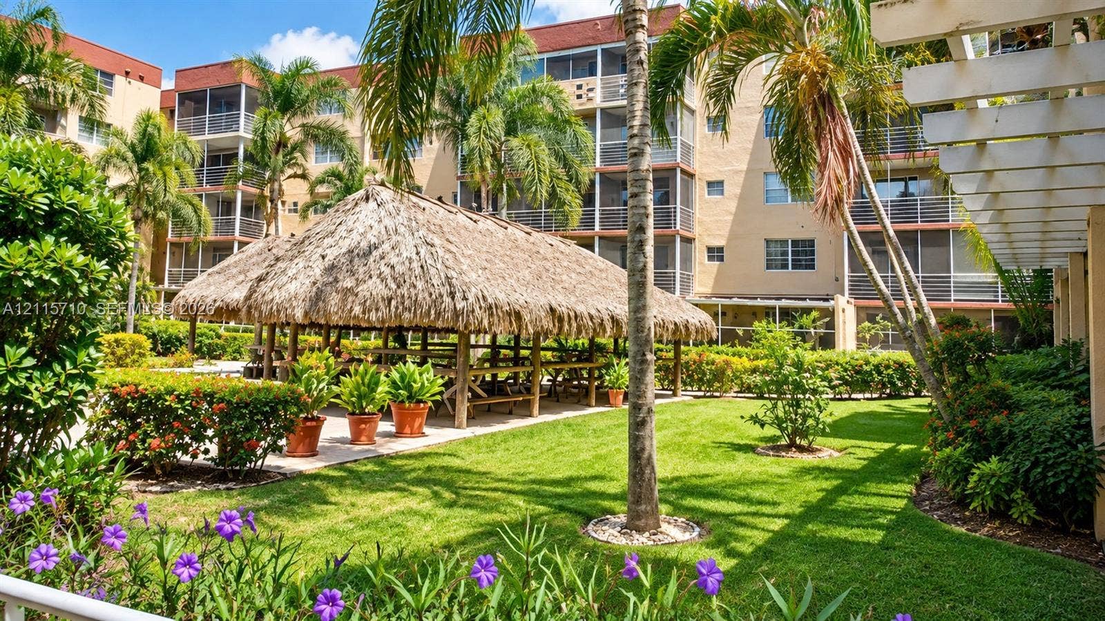406 NW 68th Avenue, #115, Plantation, Plantation, FL 33317 Manicured grounds featuring a thatched-roof pavilion, mature palm trees, decorative flowering shrubs, and multi-story building structures with screened-in balconies