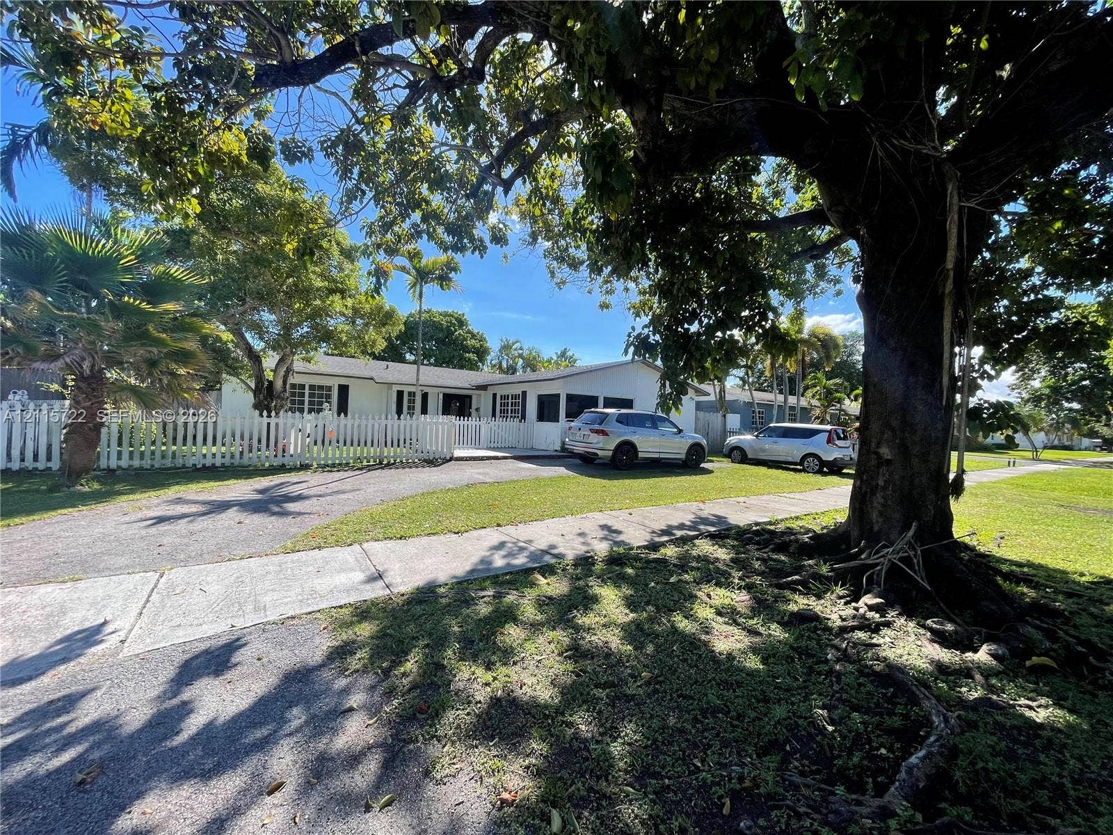 9852 SW 196th Street, Cutler Bay, FL 33157 Exterior facade featuring light-toned siding, a white picket fence, a covered porch area, and mature trees