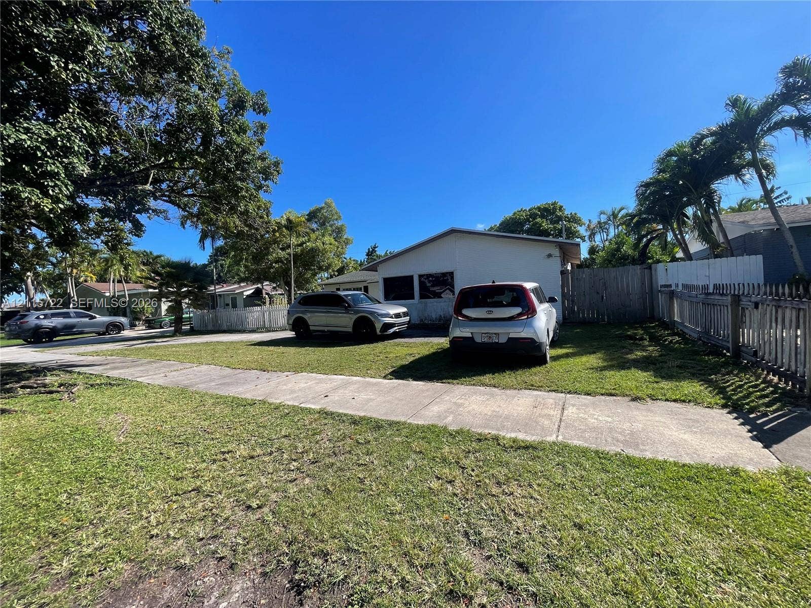9852 SW 196th Street, Cutler Bay, FL 33157 Exterior facade featuring a concrete driveway, grassy lawn, mature trees, and wooden perimeter fencing