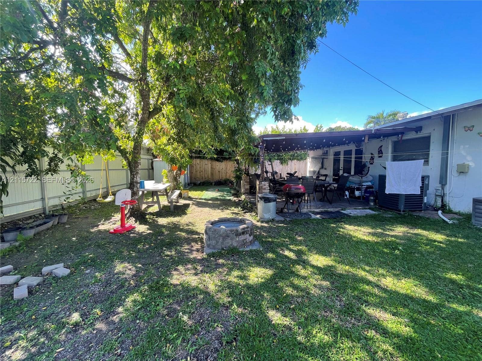9852 SW 196th Street, Cutler Bay, FL 33157 Backyard featuring a stone-encased fire pit, covered patio, mature canopy trees, and perimeter fencing