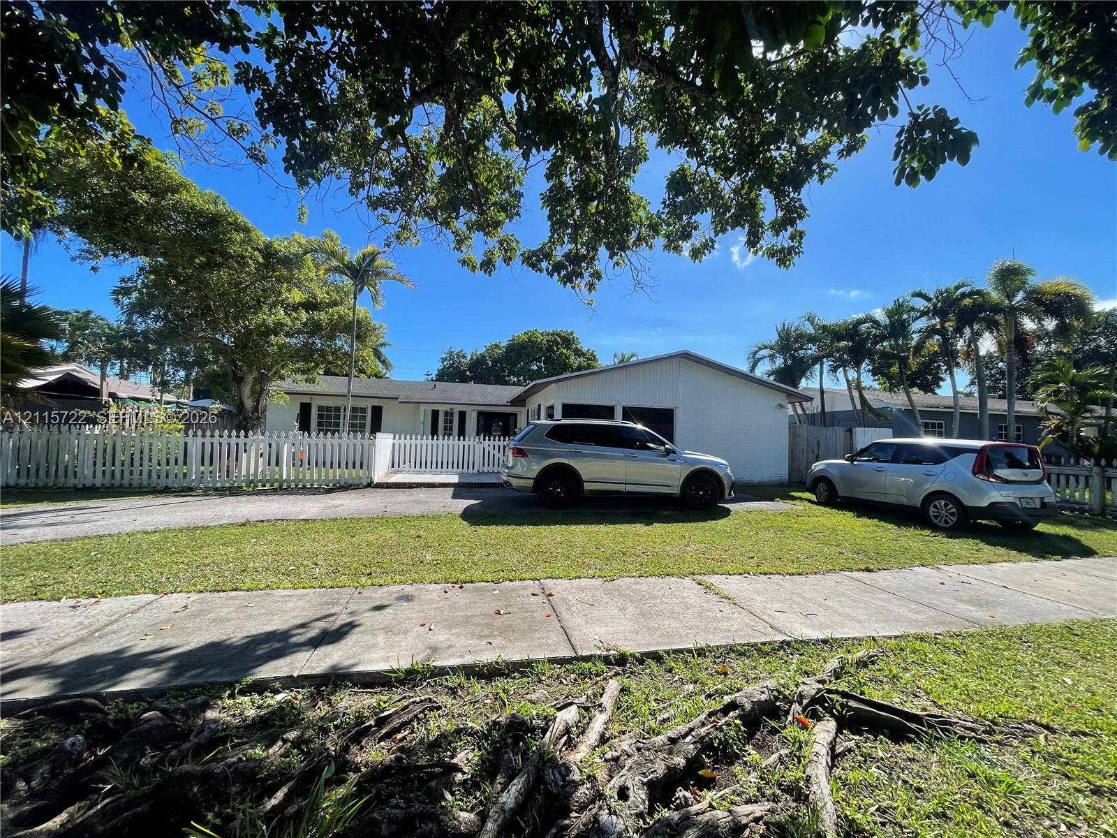 9852 SW 196th Street, Cutler Bay, FL 33157 Single-story residence featuring a light-colored facade, white picket fencing, an asphalt driveway, and mature trees