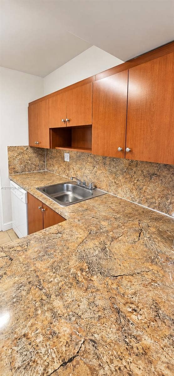 11110 SW 196th Street, #212, Cutler Bay, Cutler Bay, FL 33157 Kitchen featuring wood-finish cabinetry, stone-patterned countertops, matching stone-patterned backsplash, stainless steel drop-in sink with a chrome faucet, and a white built-in dishwasher