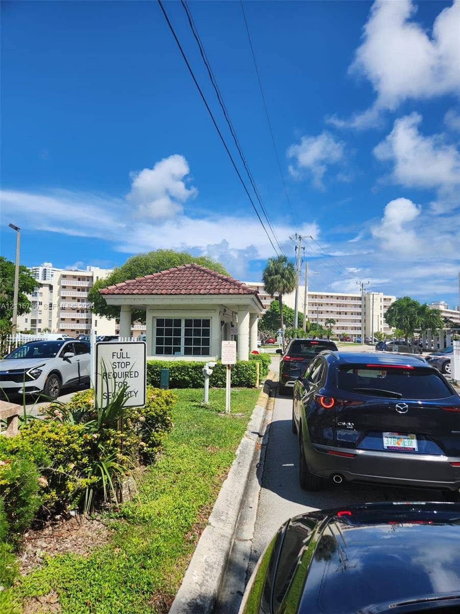 2861 Leonard Drive, #F605, Aventura, FL 33160 Gated community entrance featuring a guardhouse with a tiled roof and landscaped perimeter