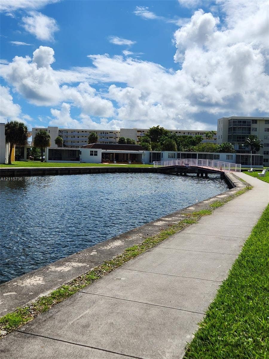 2861 Leonard Drive, #F605, Aventura, FL 33160 Waterfront grounds featuring a concrete walking path, a pedestrian footbridge, manicured lawn, and mature palm trees