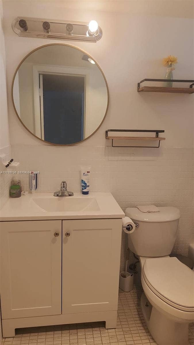 2861 Leonard Drive, #F605, Aventura, FL 33160 White vanity with integrated rectangular sink, round gold-rimmed mirror, multi-bulb wall-mounted fixture, and dual wall-mounted floating wood shelves