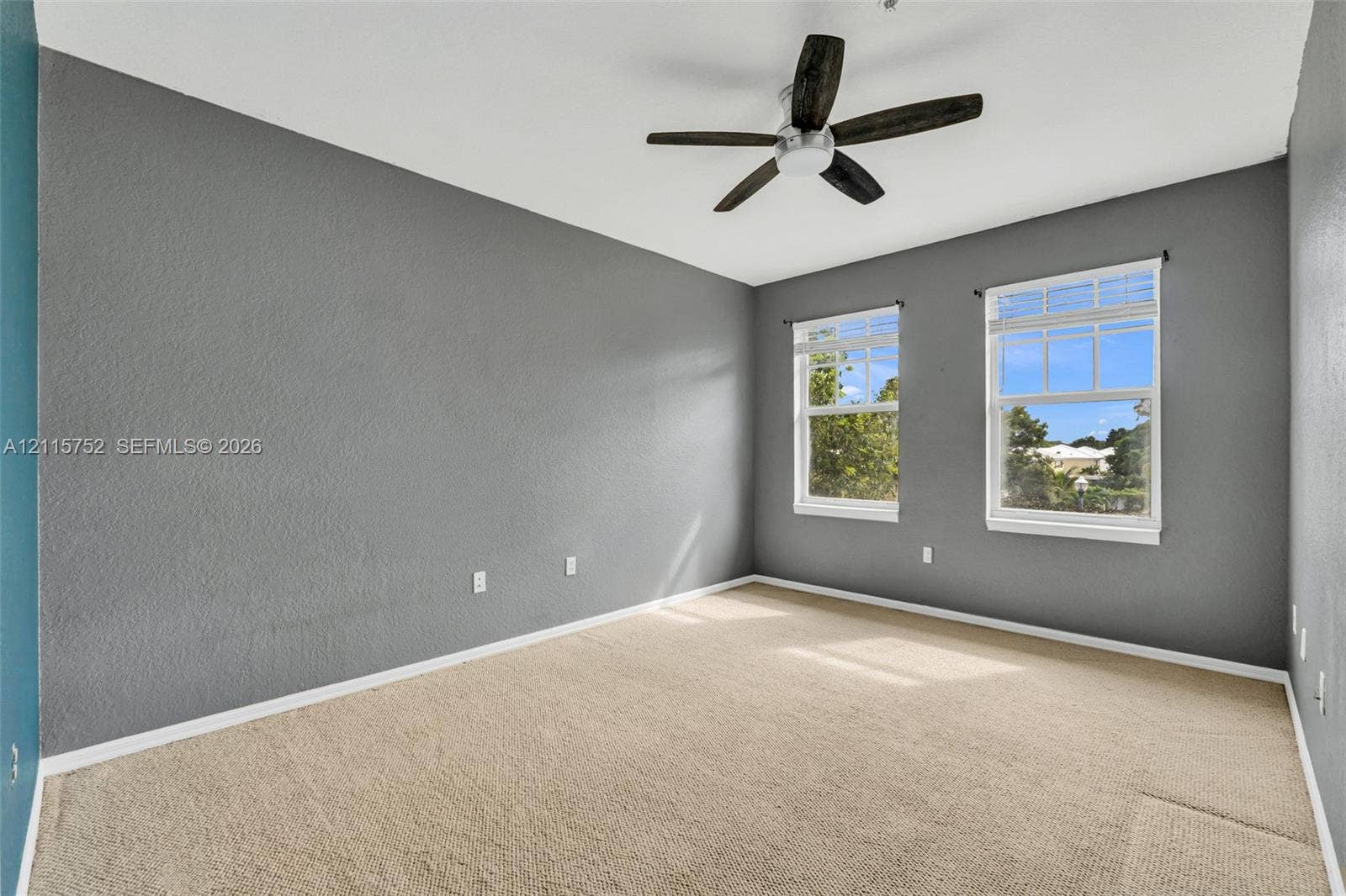 Room featuring neutral wall-to-wall carpeting, grey textured walls, dual windows, and a multi-blade ceiling fan