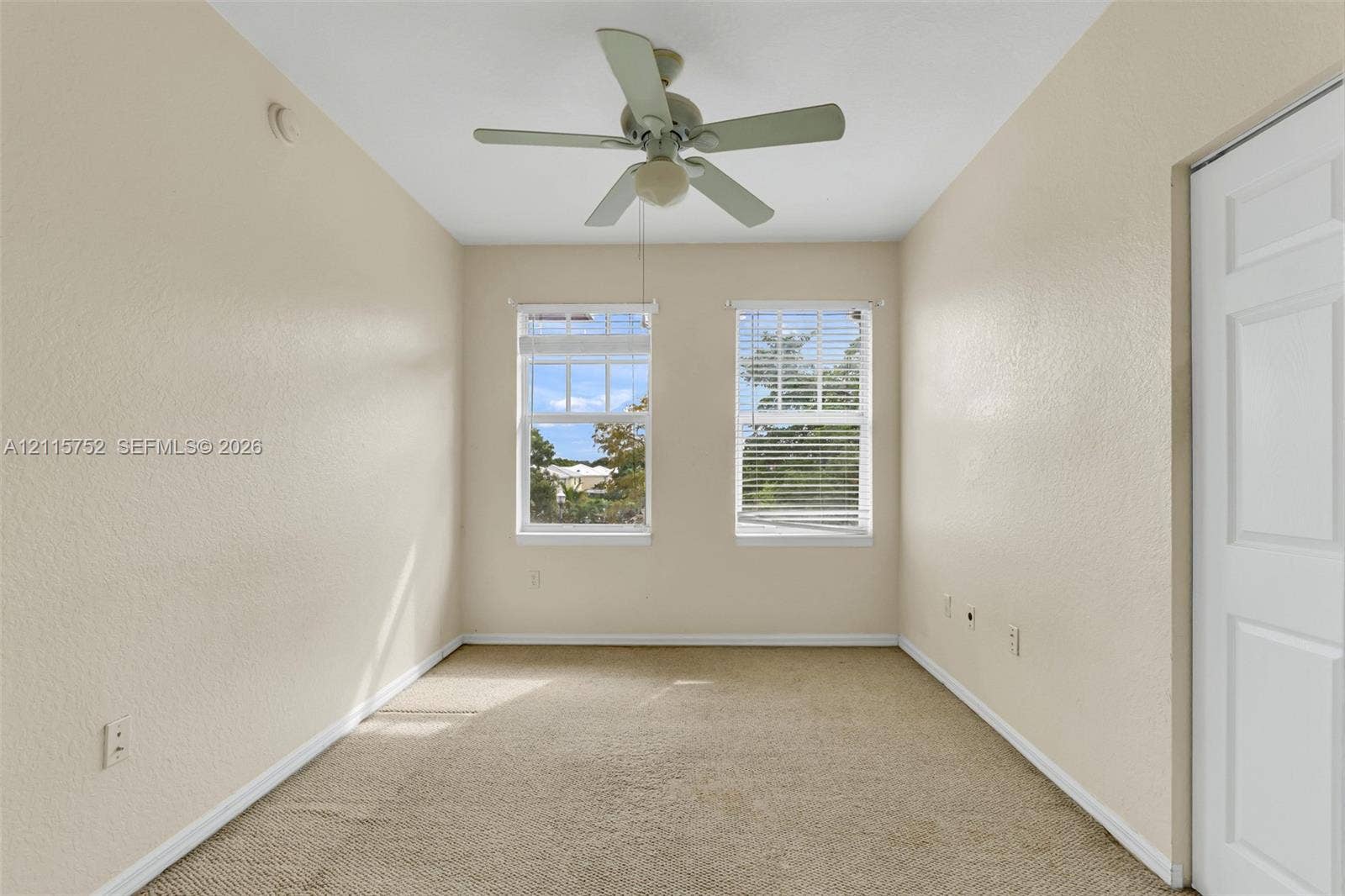 Neutral-toned interior space featuring carpeting, dual windows with blinds, a ceiling fan with integrated lighting, and a paneled closet door