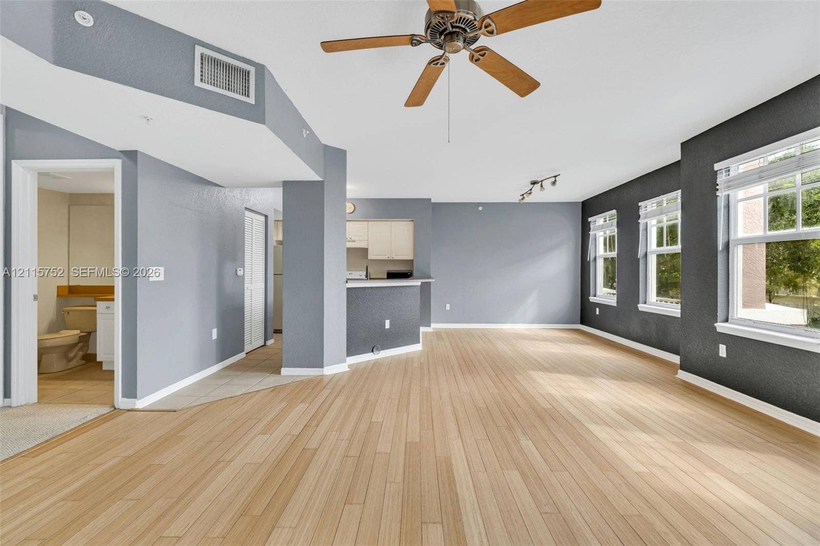Open-concept interior featuring light wood-finish flooring, a neutral color palette, three large windows with horizontal blinds, and a ceiling fan with integrated lighting