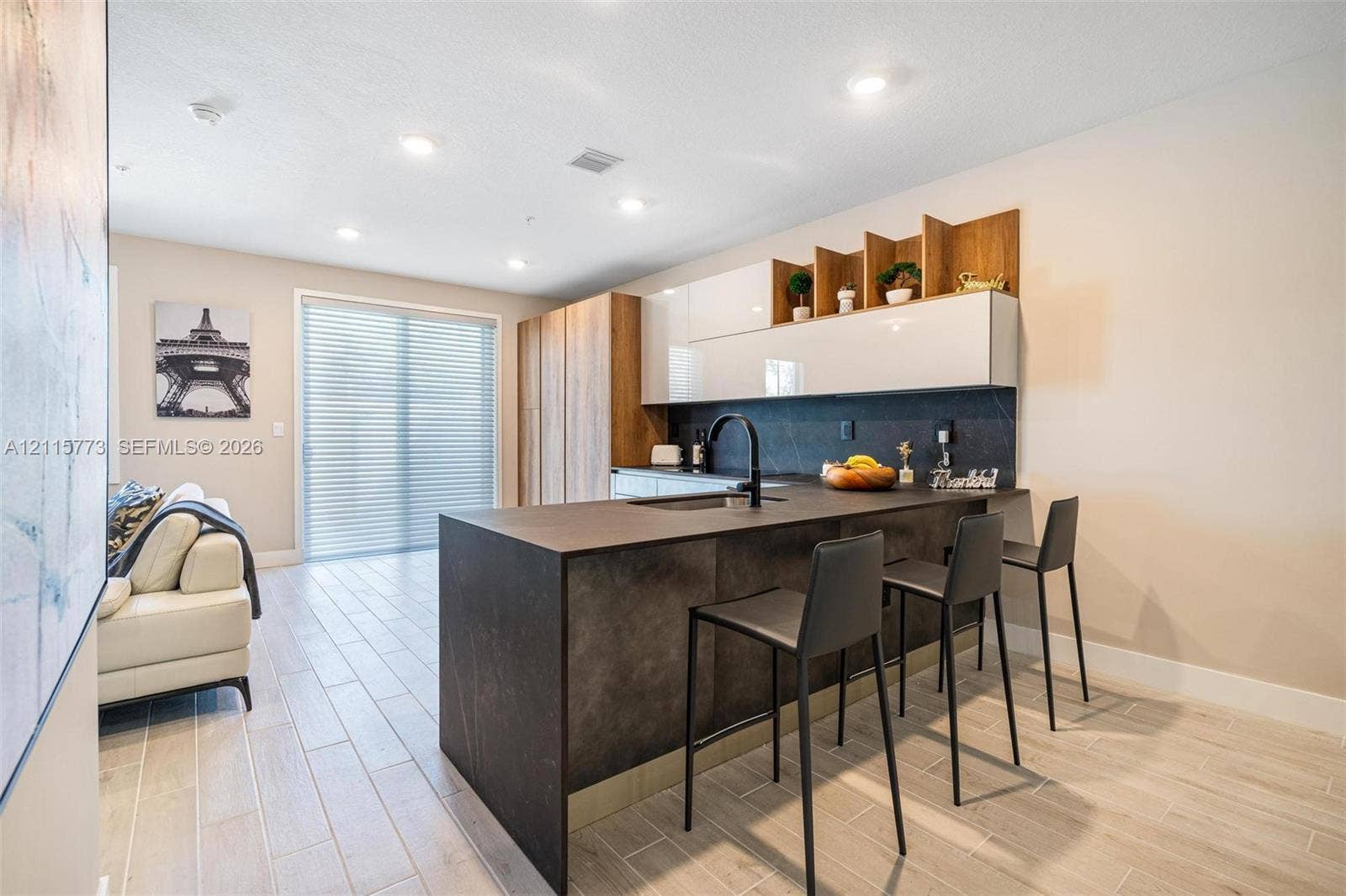 17033 SW 1st Street, #1, Pembroke Pines, Pembroke Pines, FL 33027 Kitchen featuring a matte dark finish island with a built-in sink, high-gloss white cabinetry, integrated wood-tone shelving, stone-finish backsplash, and light-toned wood-finish flooring