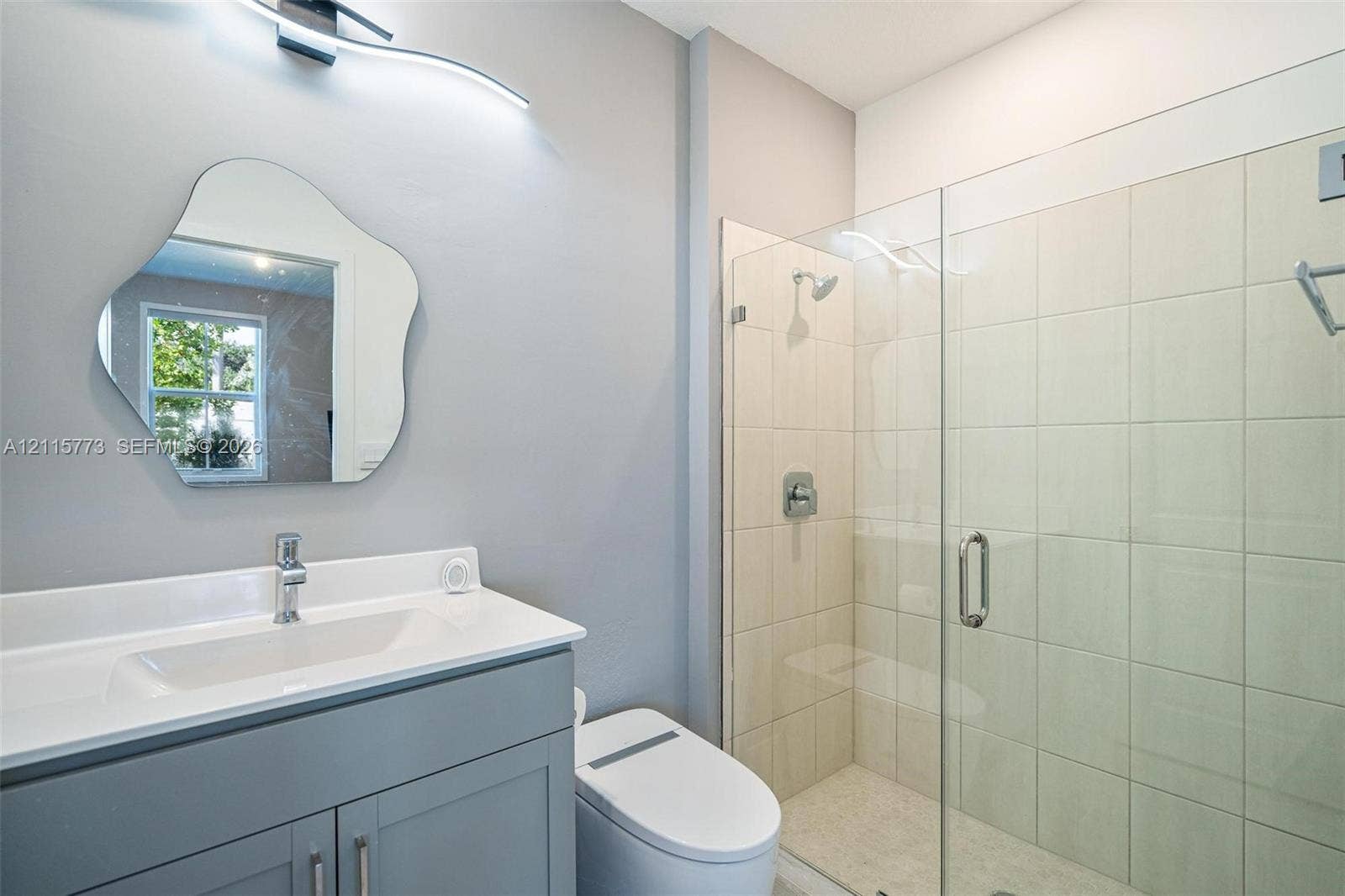 17033 SW 1st Street, #1, Pembroke Pines, Pembroke Pines, FL 33027 Grey vanity with a white countertop and integrated sink, chrome faucet, wall-mounted curved light fixture, organic-shaped mirror, and walk-in shower with a glass enclosure and tiled surround