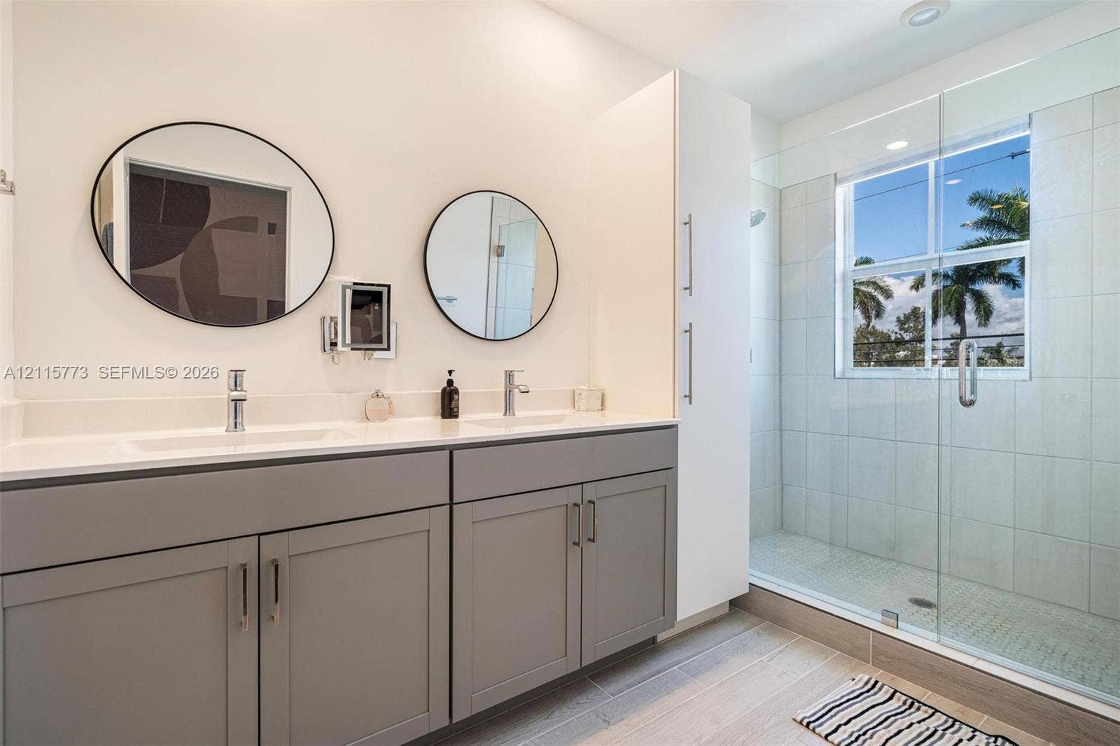 17033 SW 1st Street, #1, Pembroke Pines, Pembroke Pines, FL 33027 Dual-sink vanity with light-colored countertops, grey cabinetry, wall-mounted circular mirrors, a tall floor-to-ceiling storage cabinet, and a walk-in shower with glass enclosure and light-toned tile surround
