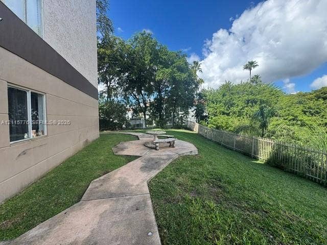 Ground-level exterior area featuring a concrete walkway, a stone-finish circular table with benches, manicured lawn, and perimeter metal fencing