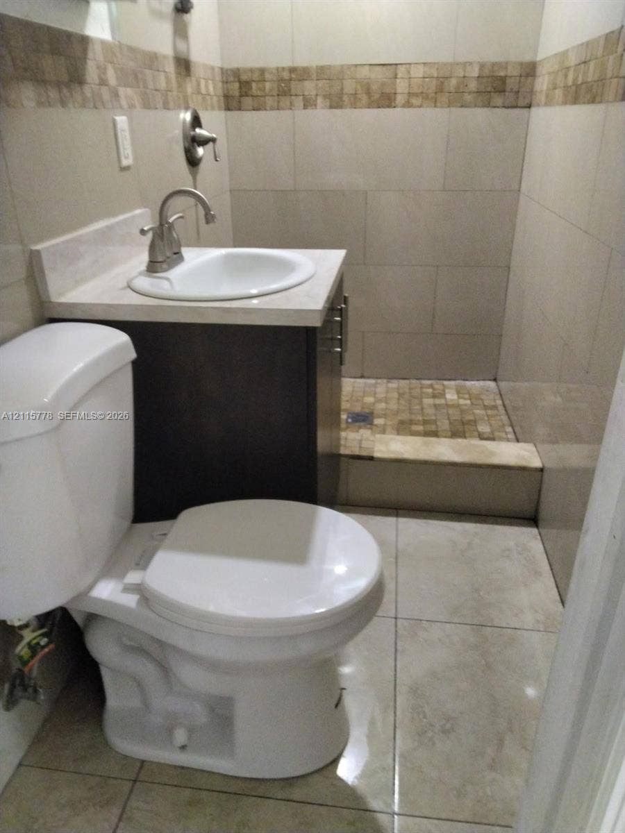 Bathroom featuring a dark wood-finish vanity with a stone-top integrated sink, a tiled walk-in shower with a mosaic accent border, and large-format neutral floor tiling