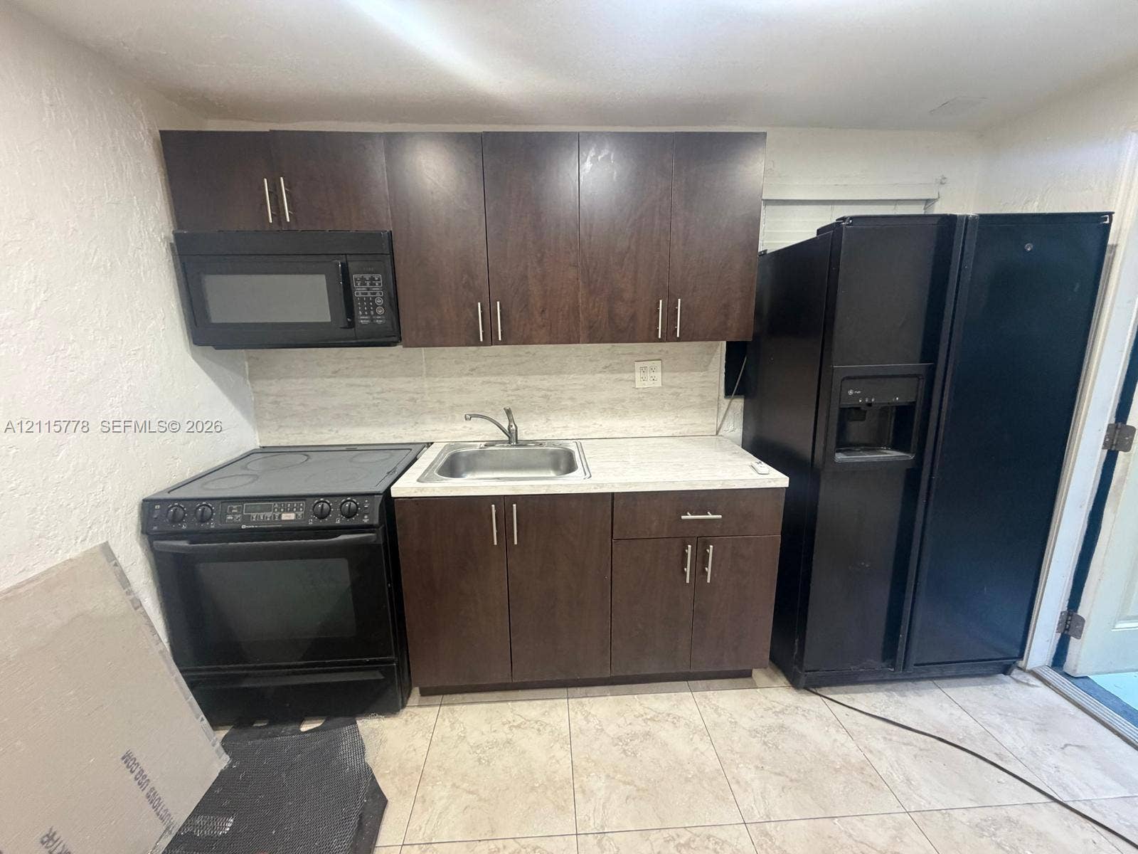 Dark wood-finish cabinetry, stone-surface countertop, stainless steel sink with a high-arch faucet, tile flooring, and integrated black kitchen appliances