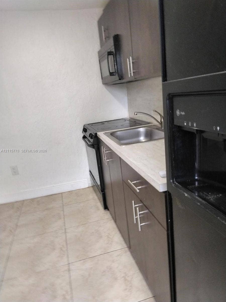 Kitchenette featuring dark-finish cabinetry with modern hardware, neutral stone-finish countertops, stainless steel sink with gooseneck faucet, built-in microwave, and tile flooring