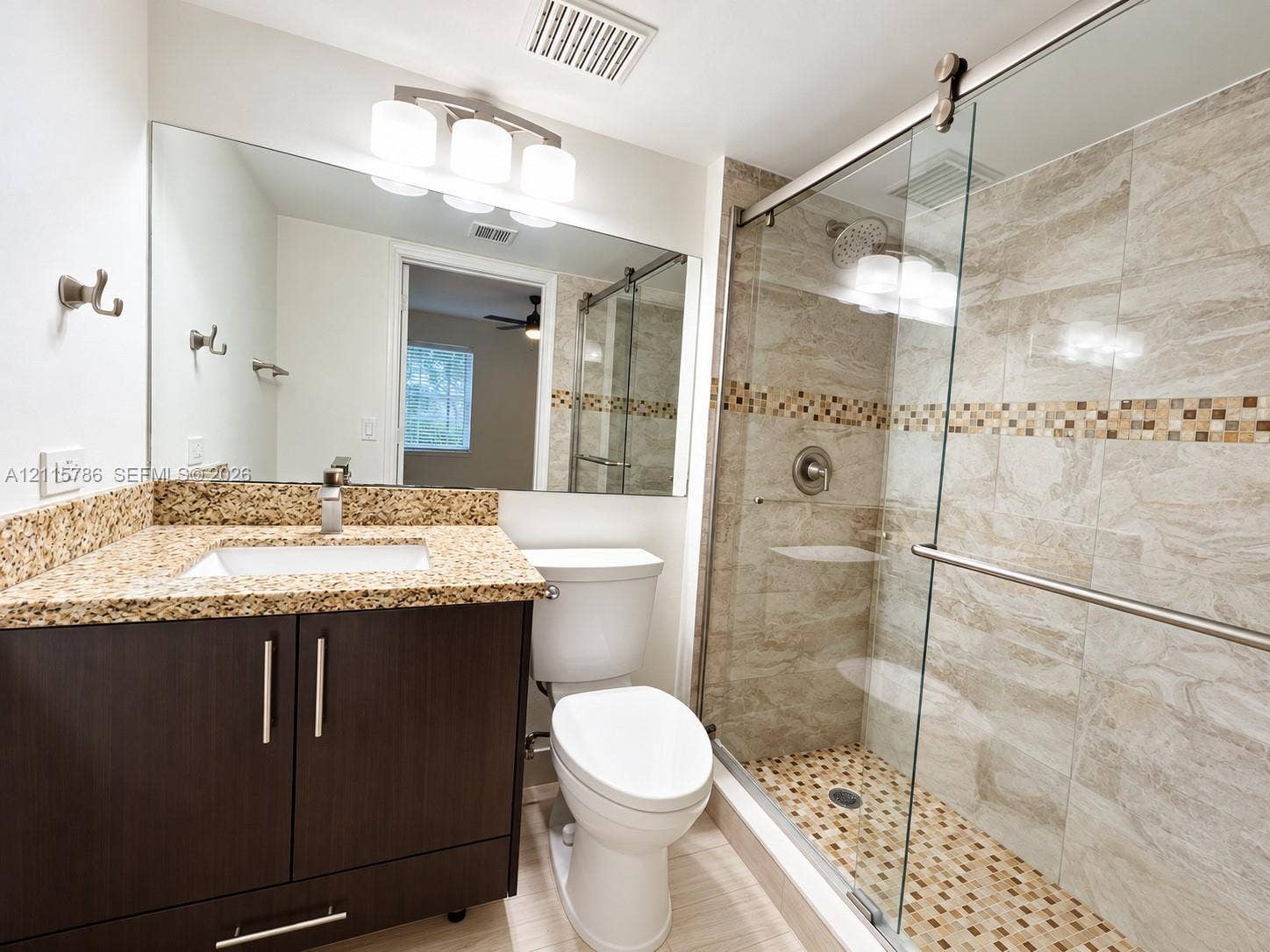 2494 Centergate Drive, #202, Miramar, Miramar, FL 33025 Dark wood-finish vanity with stone-composite countertop, full-width vanity mirror, triple-bulb vanity light, and a walk-in shower featuring glass sliding doors and decorative tile inlay