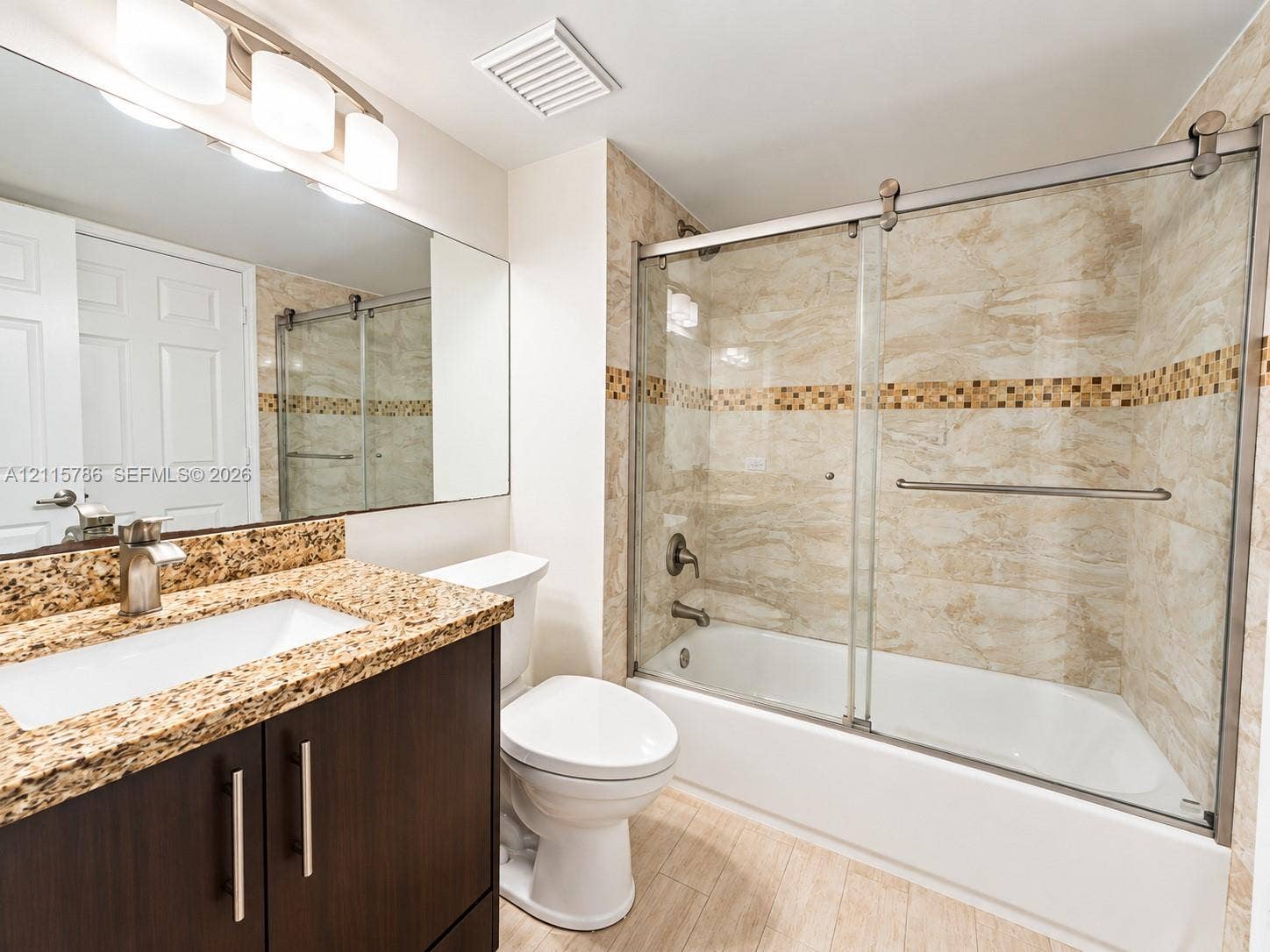 2494 Centergate Drive, #202, Miramar, Miramar, FL 33025 Dark wood-finish cabinetry, stone countertop with an integrated rectangular basin, full-width wall mirror, and a shower-tub combination with sliding glass doors and decorative tile inlay