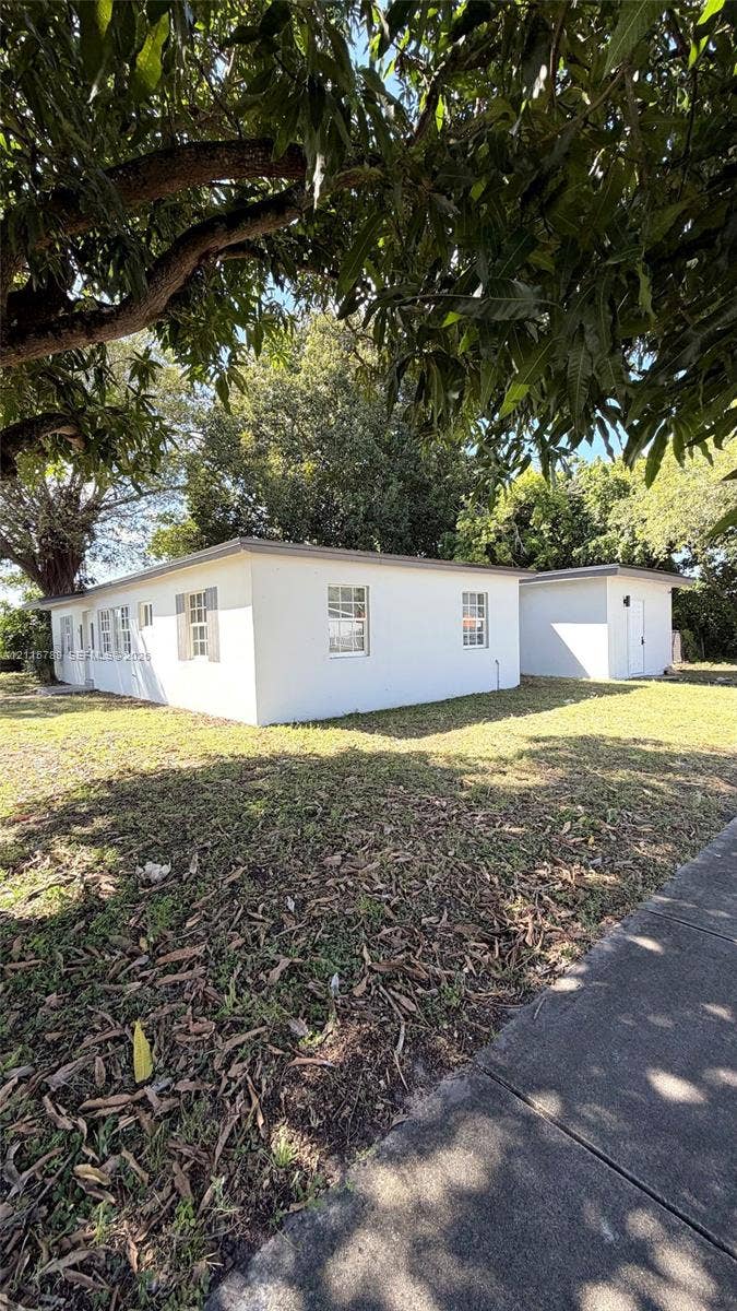 16220 NW 28th Place, Miami Gardens, FL 33054 Stucco exterior featuring multi-pane windows with decorative shutters, flat roofline, and an auxiliary structure situated on a lot with mature trees