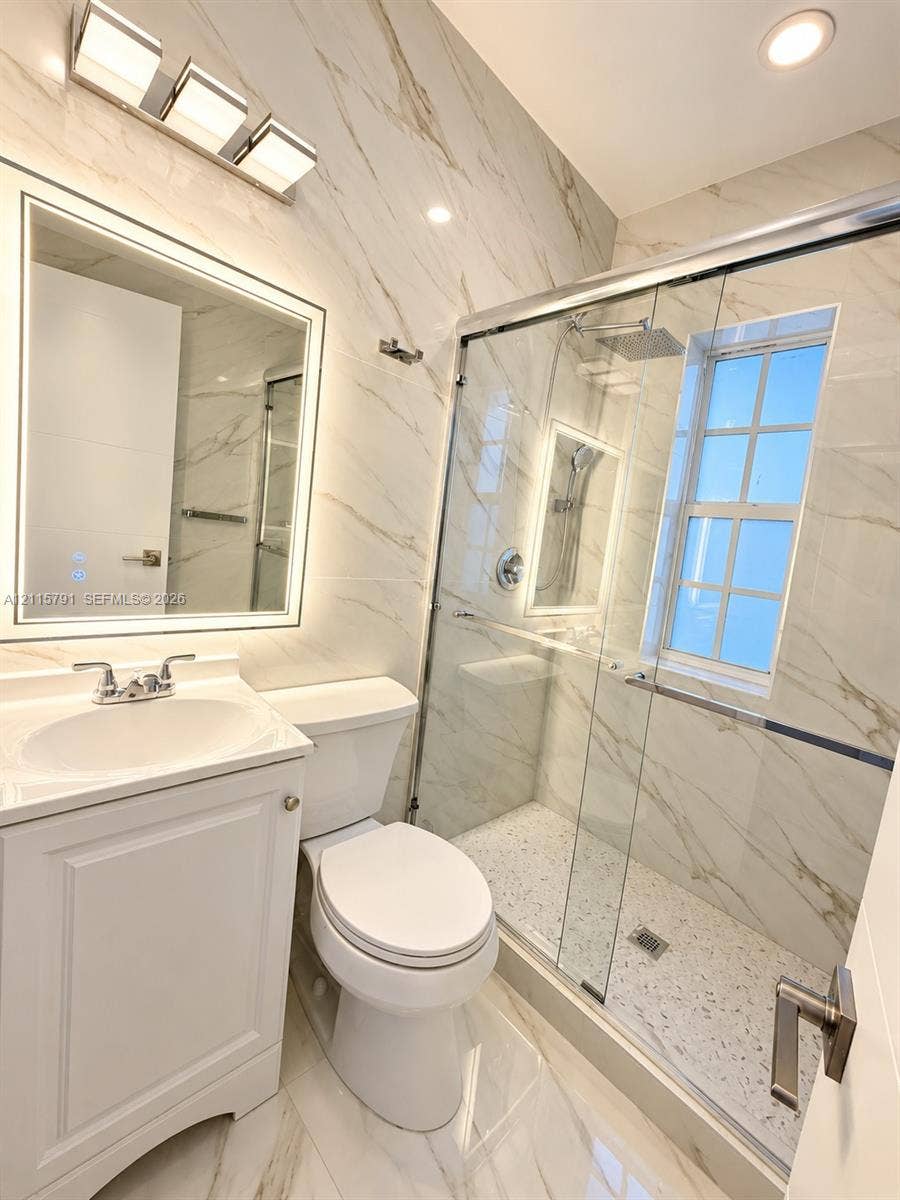 1455 Michigan Avenue, #19B, Miami Beach, Miami Beach, FL 33139 Bathroom featuring full-height stone-finish wall tiling, a glass-enclosed shower with a rainfall showerhead, a backlit vanity mirror, and white shaker-style cabinetry