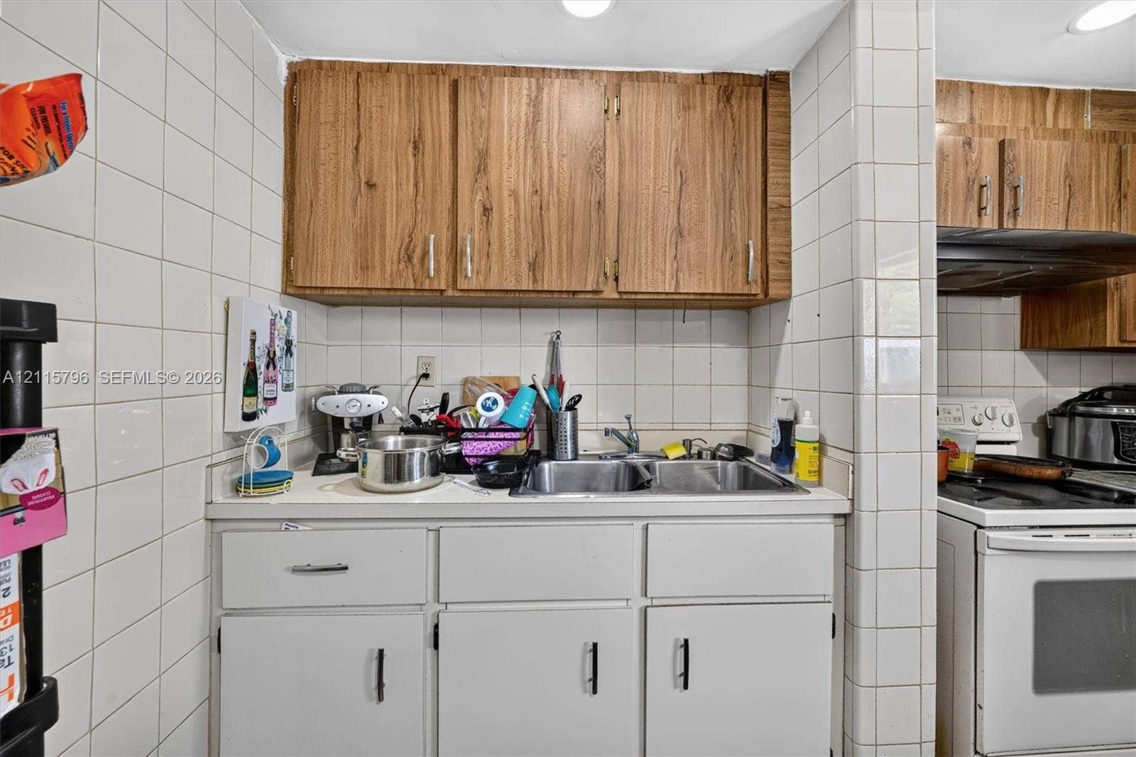 Kitchen area featuring wood-finish upper cabinetry, white lower cabinets, square tile backsplash, and dual-basin stainless steel sink