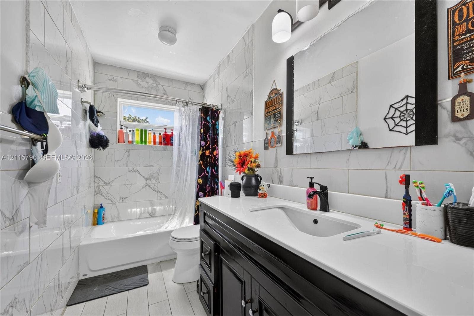 Bathroom featuring a black vanity with a white countertop and integrated sink, a deep soaking tub with a tiled surround, light-toned tiled flooring, and a large wall-mounted mirror
