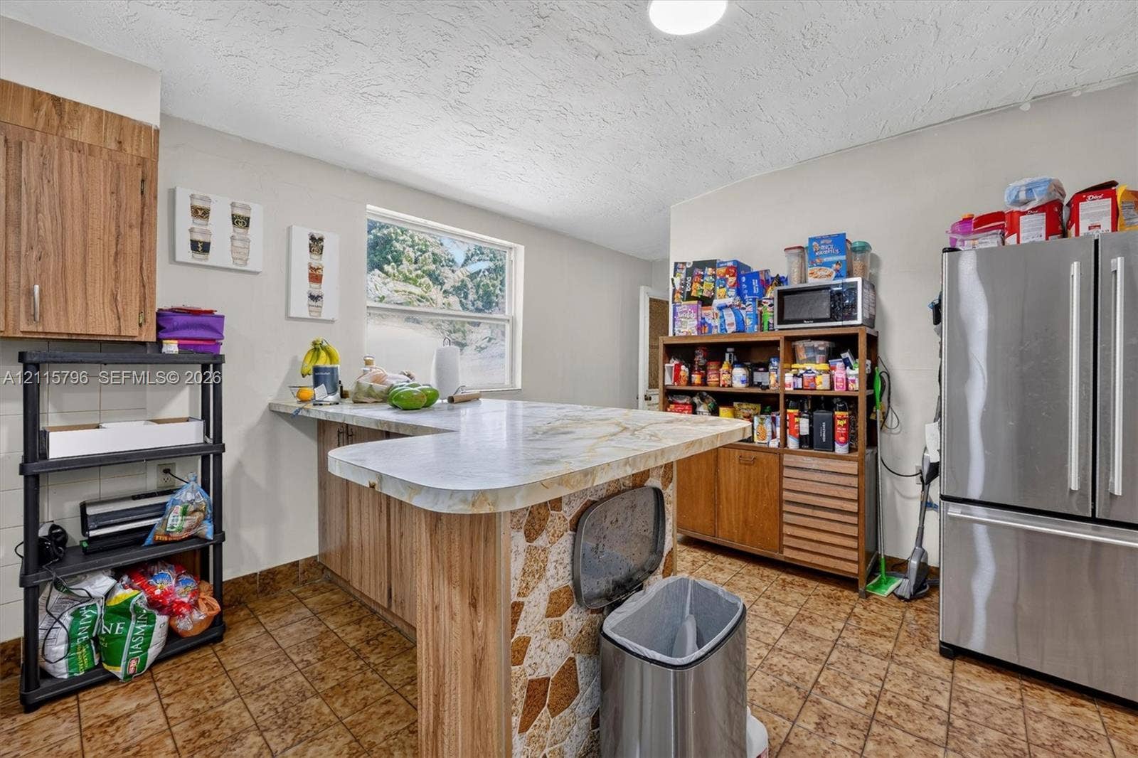 Kitchen featuring an L-shaped peninsula with a natural stone-style countertop, a multi-tone decorative stone-veneer base, stainless steel French door refrigerator, and ceramic tile flooring