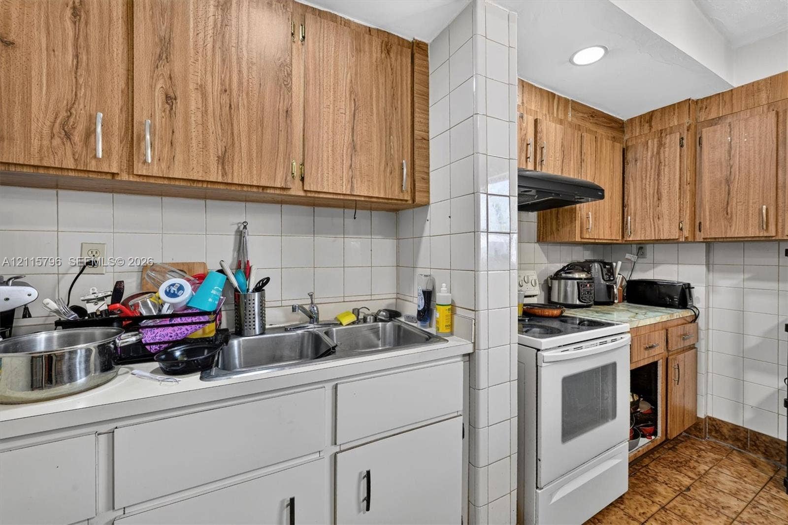 Kitchen featuring wood-finish cabinetry, white tile backsplash, double-basin stainless steel sink, and ceramic tile flooring