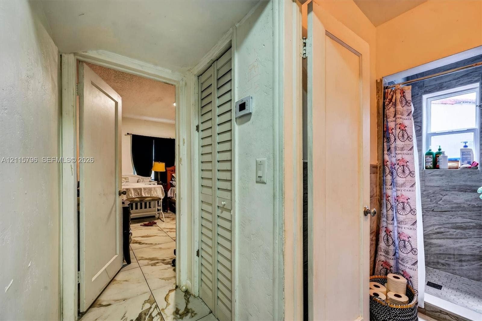 Hallway featuring louvered bi-fold closet doors, wall-mounted thermostat, and floor-to-ceiling tile shower surround with integrated window