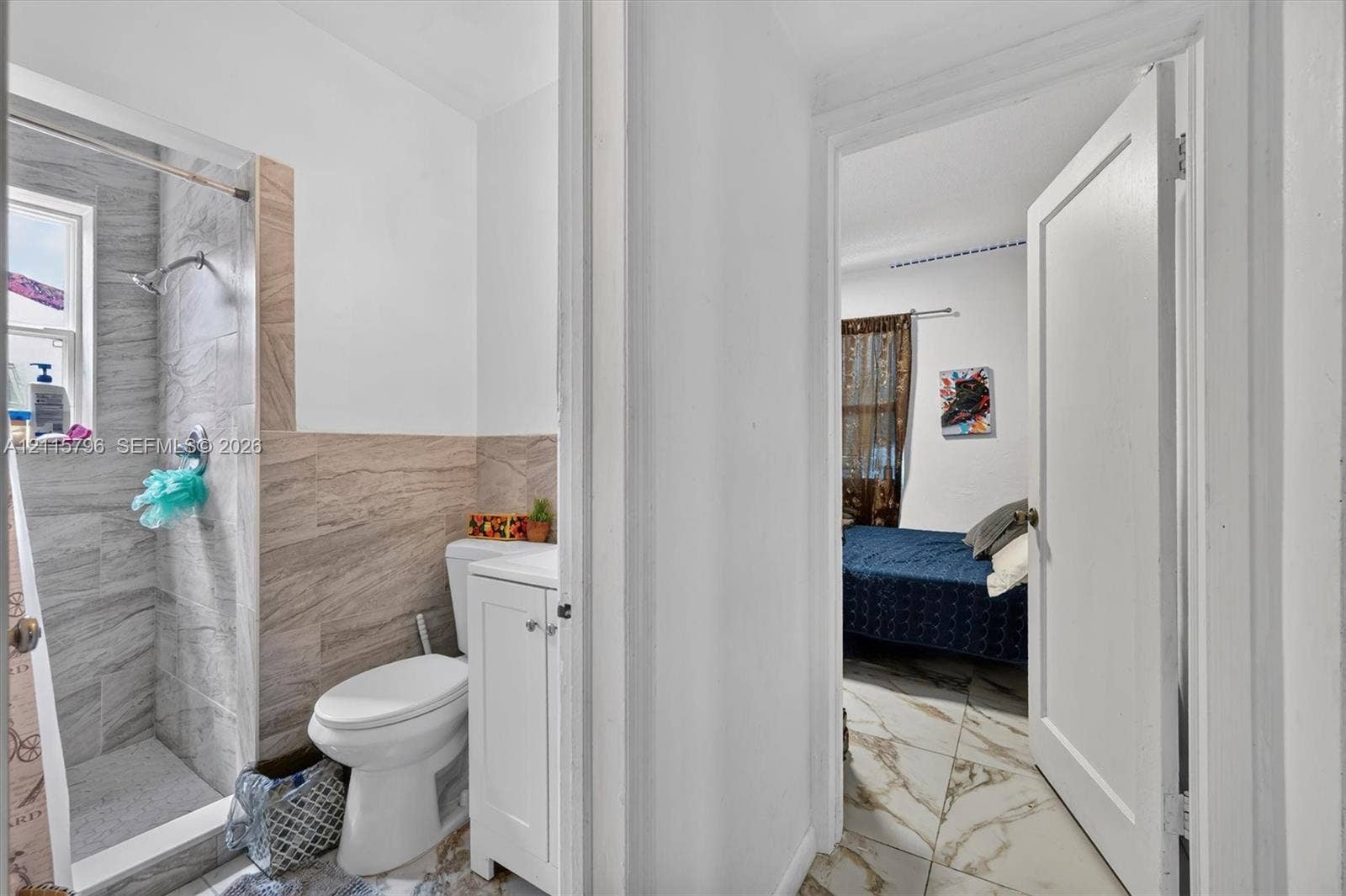 Bathroom featuring a walk-in shower with grey stone-style tile surround, beige tile wainscoting, and a white vanity with a porcelain toilet, complemented by large-format marble-finish tile flooring
