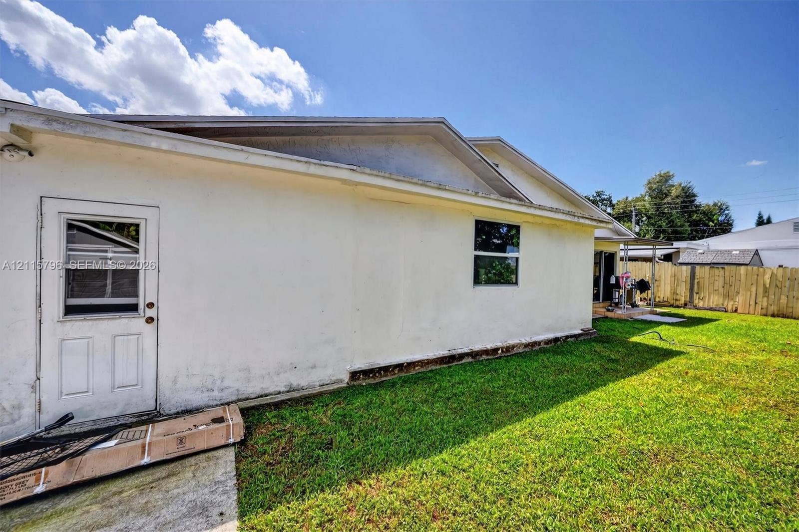 Exterior features including a light-colored stucco finish, a rear-entry door, a single-pane window, and a perimeter wood-plank fence enclosing a grass yard